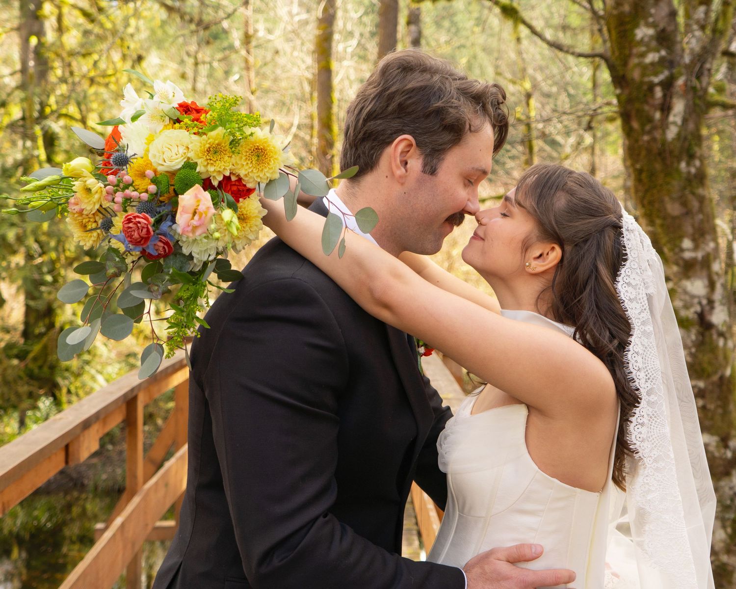 Bride and groom sharing a romantic almost-kiss moment on a wooden bridge surrounded by lush forest.