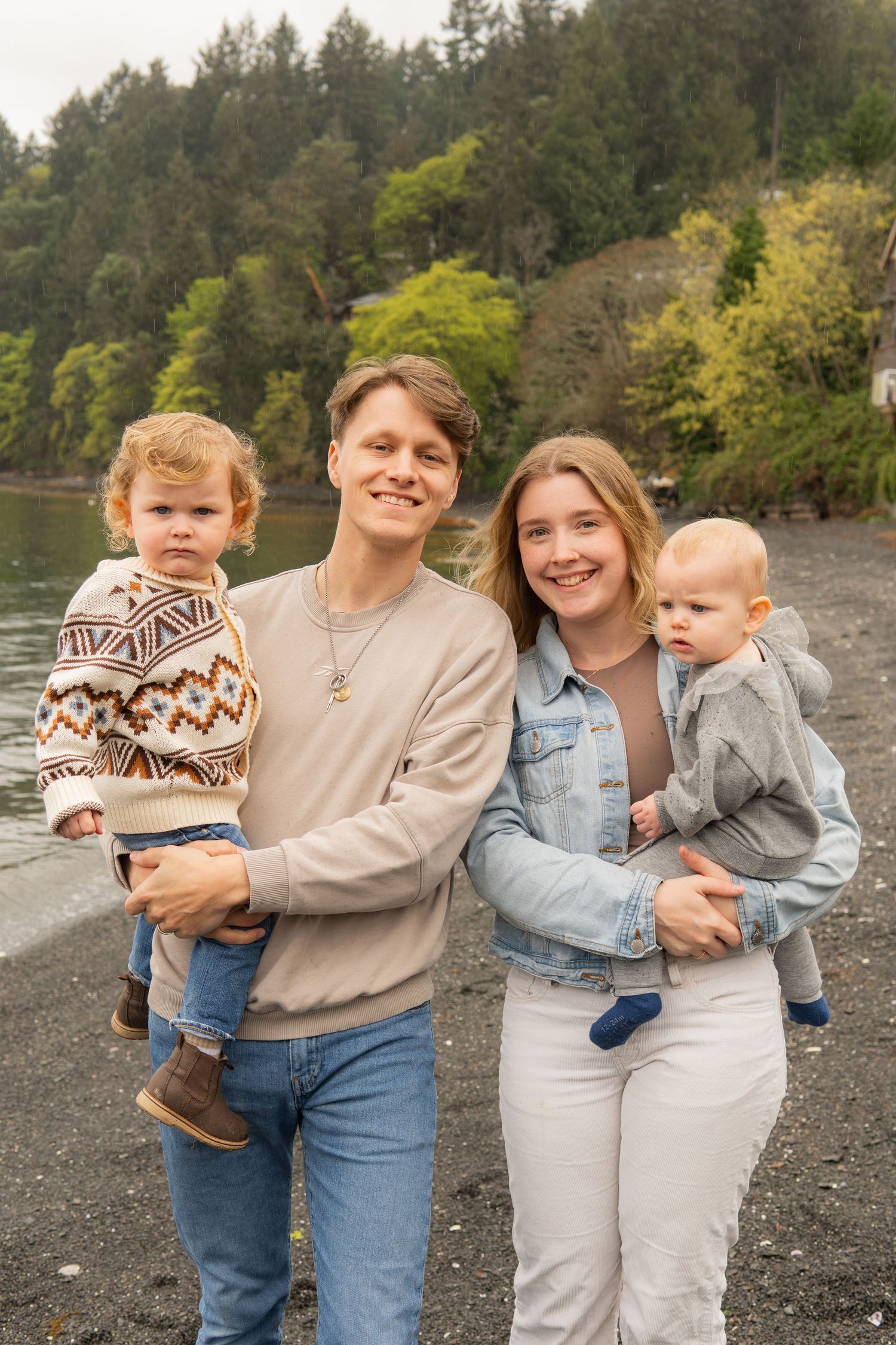Young couple smiling outdoors near a lake, each holding a toddler, with lush green trees in the background.