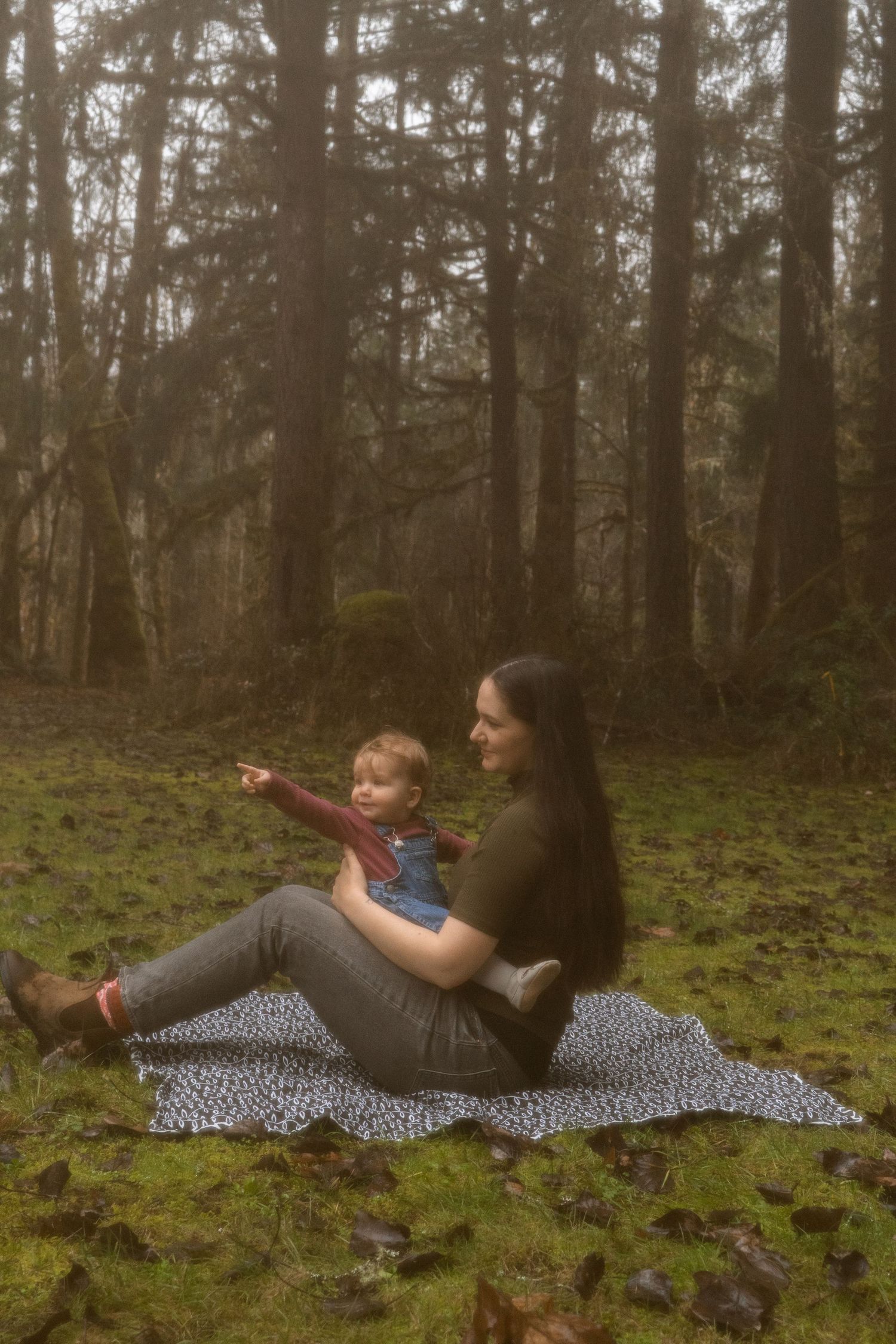 Mother and toddler sitting on blanket in forest, child pointing into the distance.