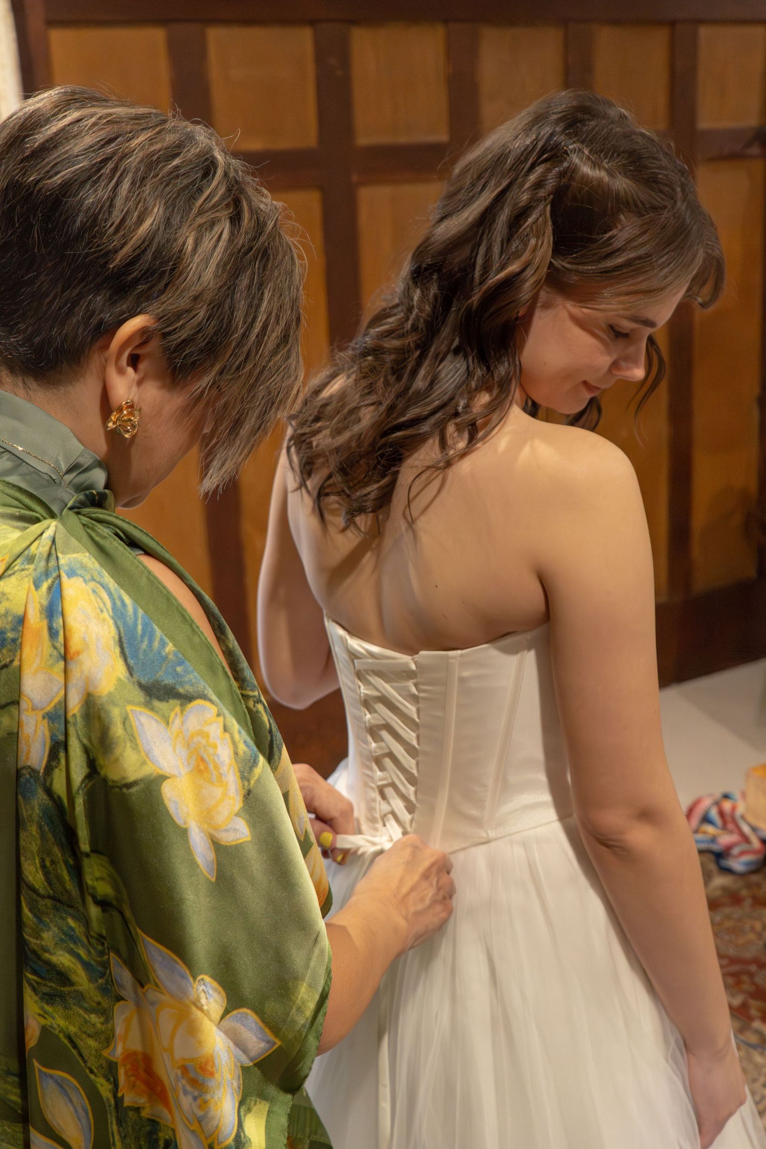 Woman in floral dress lacing up a bride's white corset wedding gown from behind.
