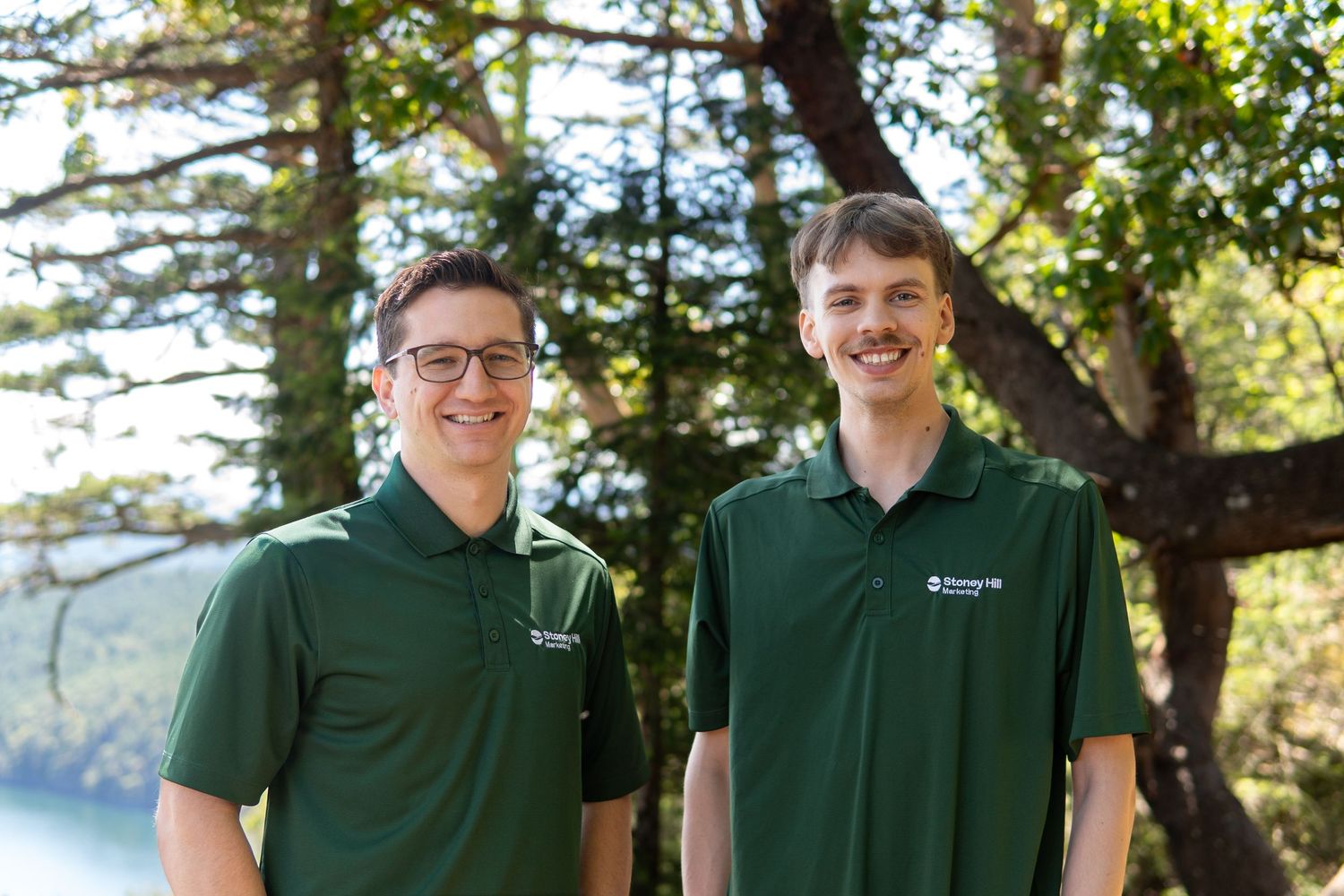 Two men in matching green polo shirts smile outdoors with trees and water in the background.