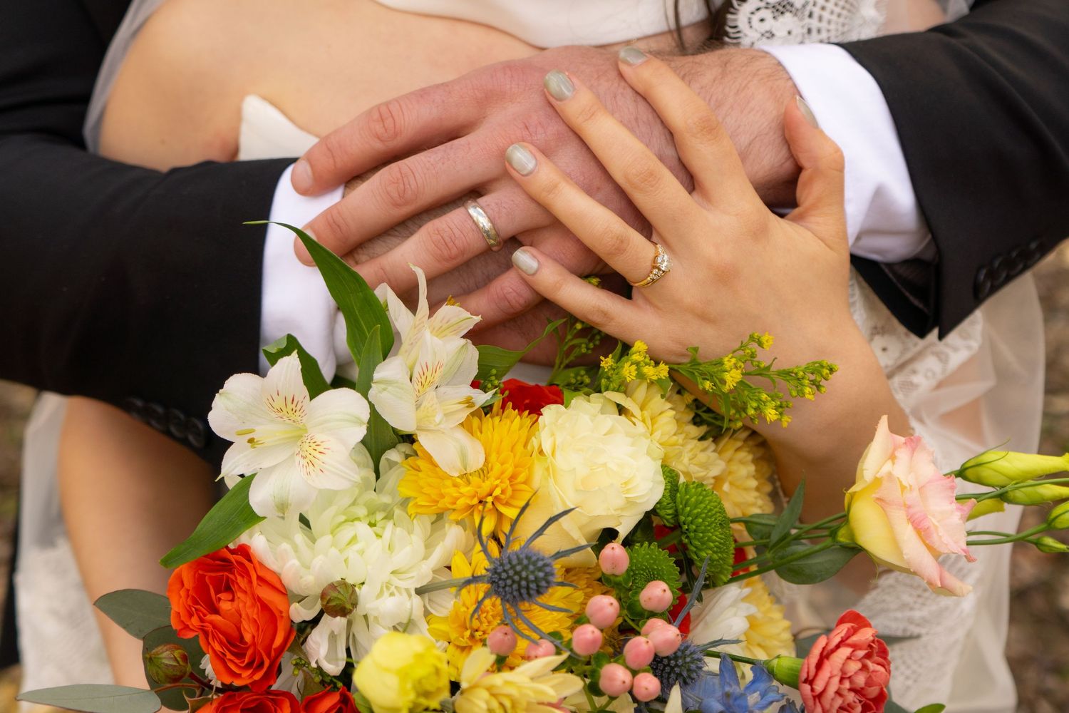 Newlyweds displaying wedding rings over a colorful bridal bouquet with white and yellow flowers.