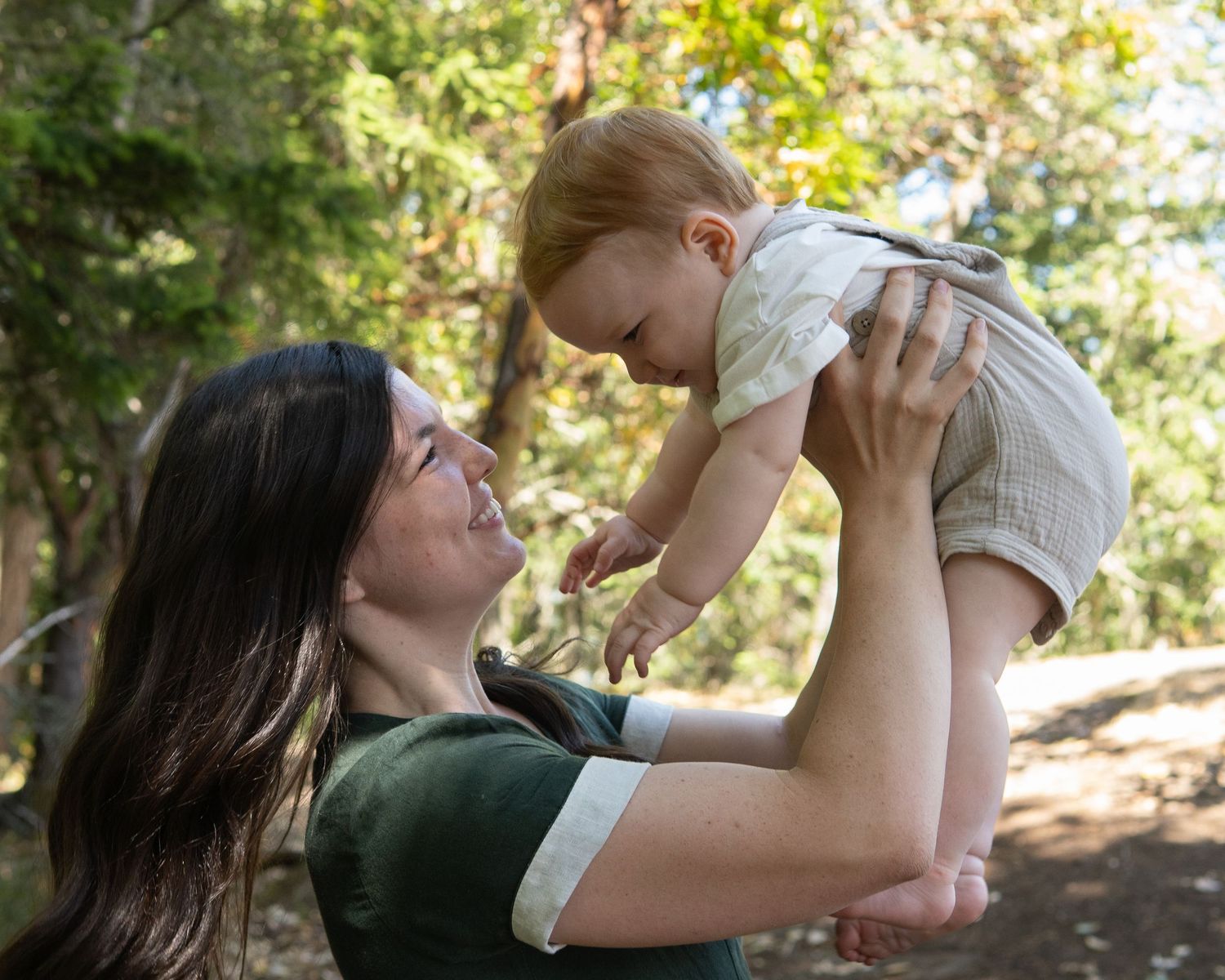 A joyful mother lifts her smiling red-haired baby in the air outdoors among green trees.