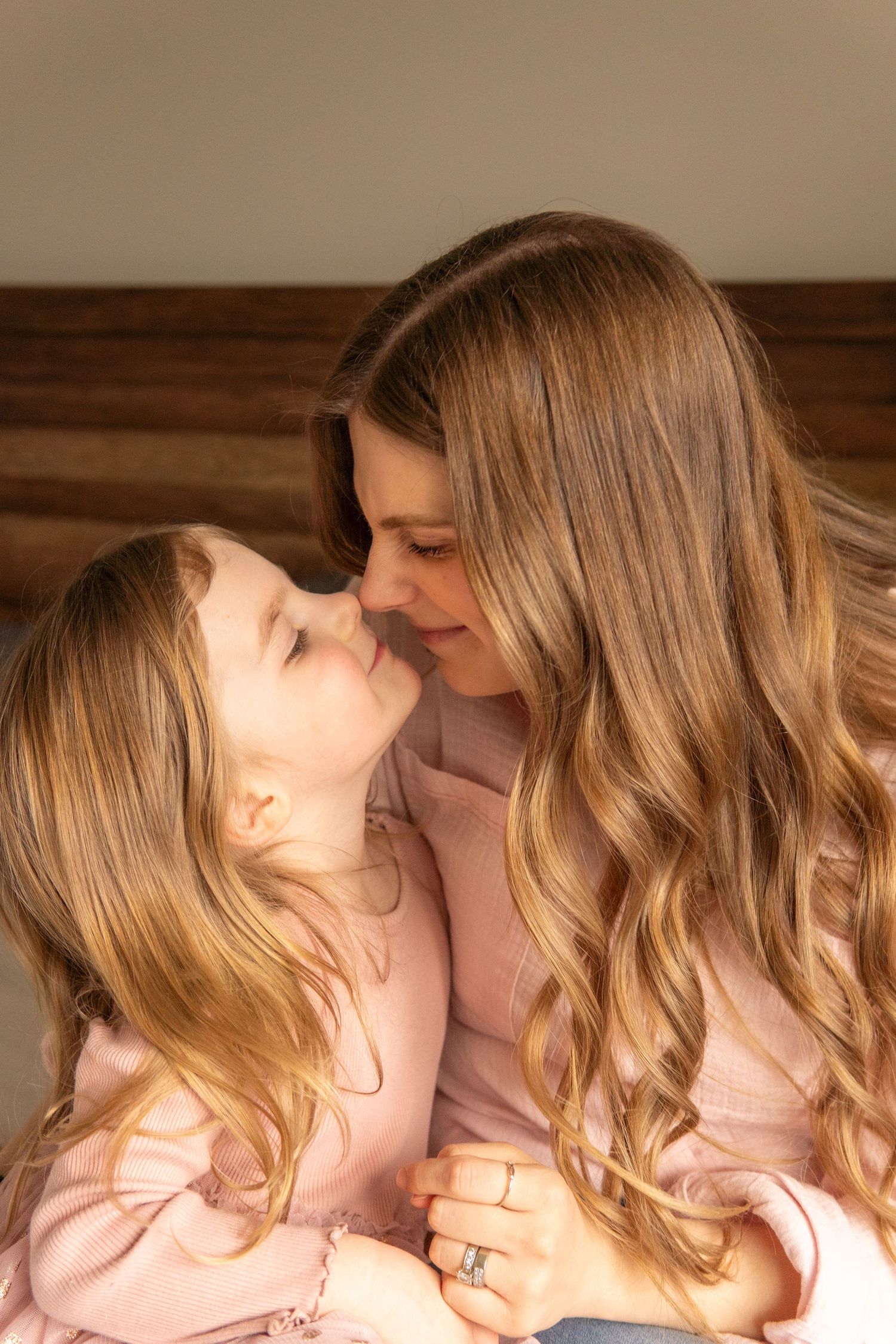 A mother and young daughter share a tender nose-to-nose moment, both wearing pink, with warm natural light.