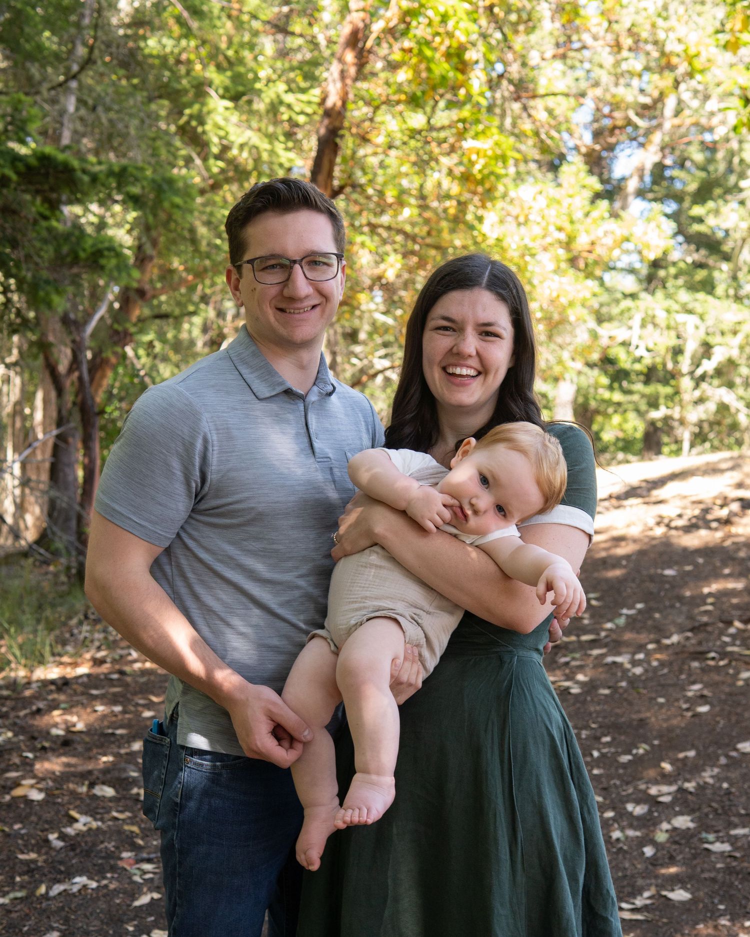 Happy young family with mother in green dress and father in gray polo holding smiling baby outdoors.