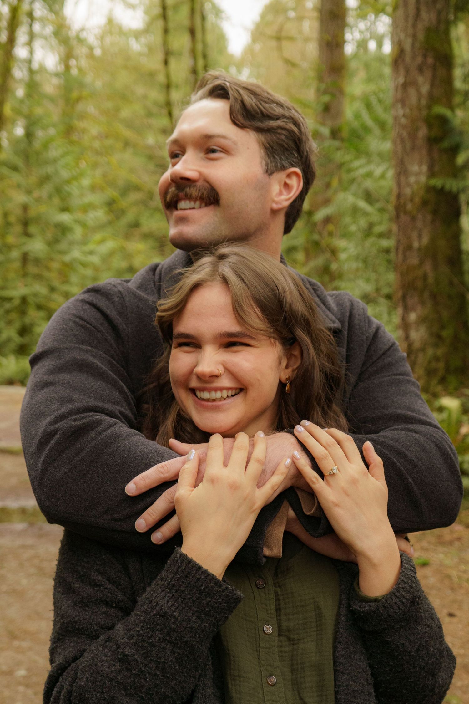 Couple embracing in a lush forest, woman smiling with engagement ring visible on her hand.