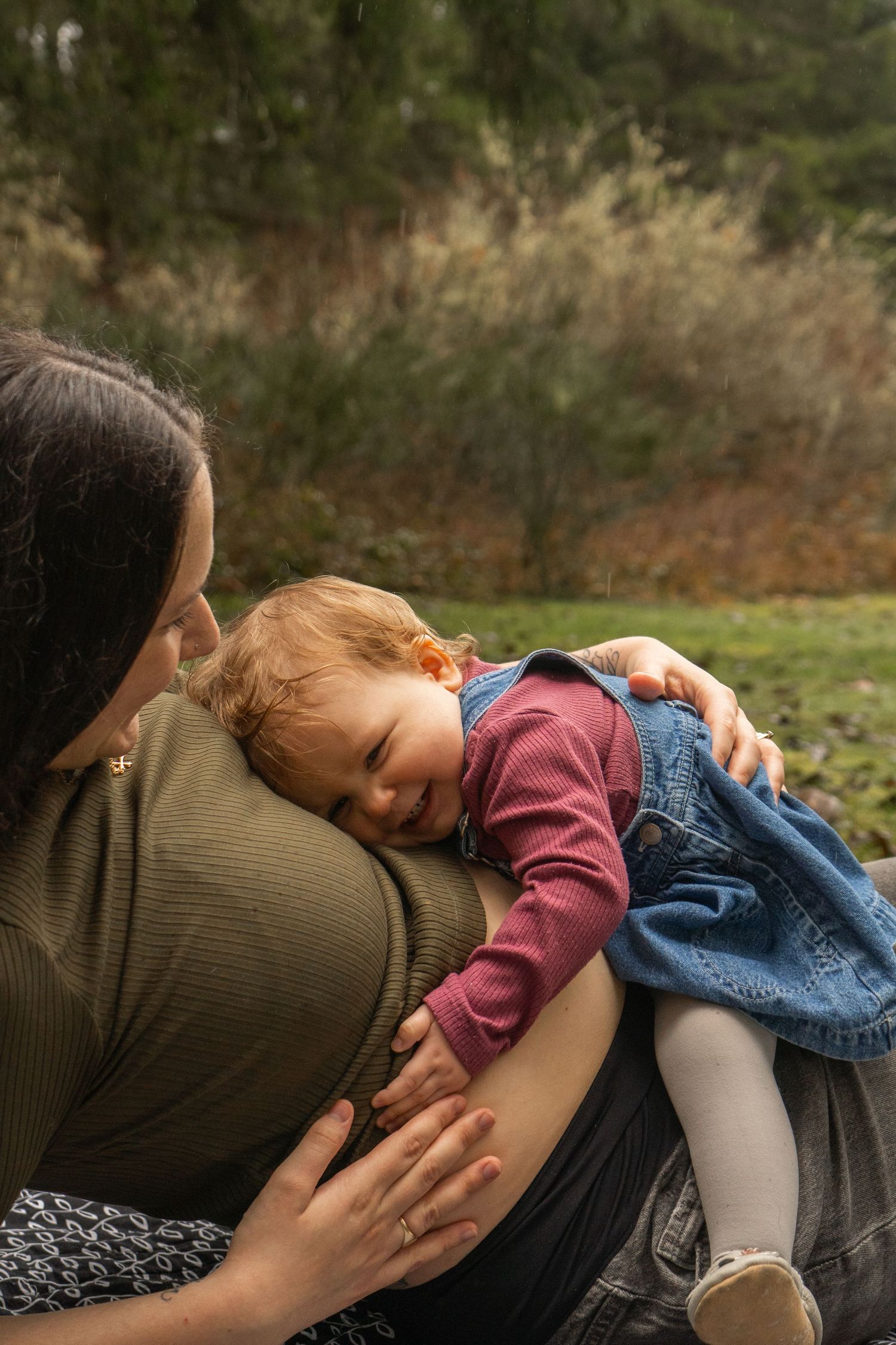 A mother tenderly holds her smiling red-haired baby outdoors, both dressed casually in a lush green setting.