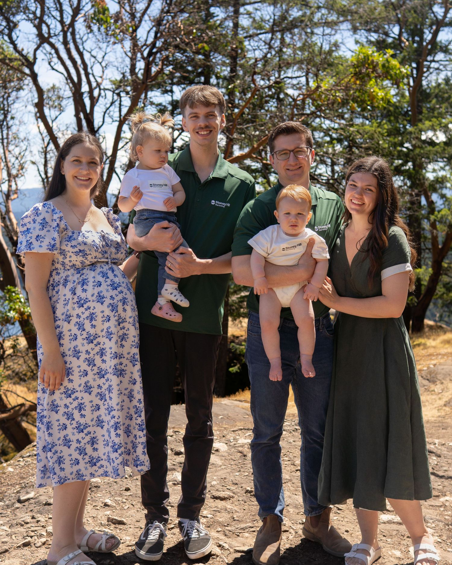 Two couples with babies pose outdoors among trees on a sunny day, smiling together in a casual family portrait.