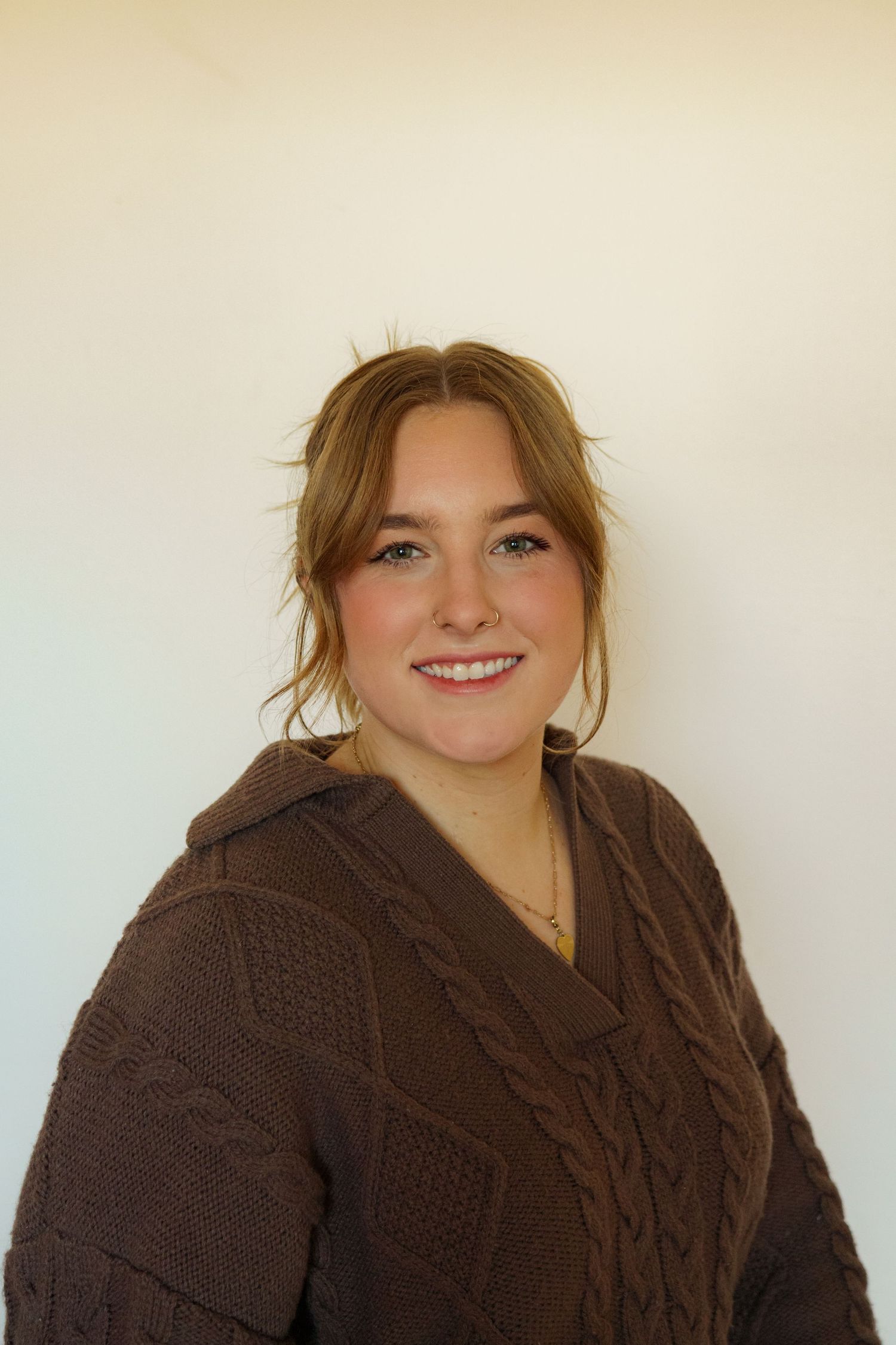 Smiling young woman with blonde hair wearing a brown cable-knit sweater posing against a white background.
