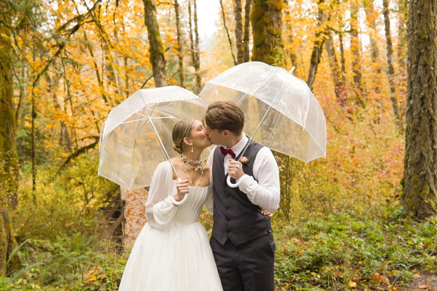 Bride and groom kissing under clear umbrellas surrounded by stunning golden autumn forest foliage.