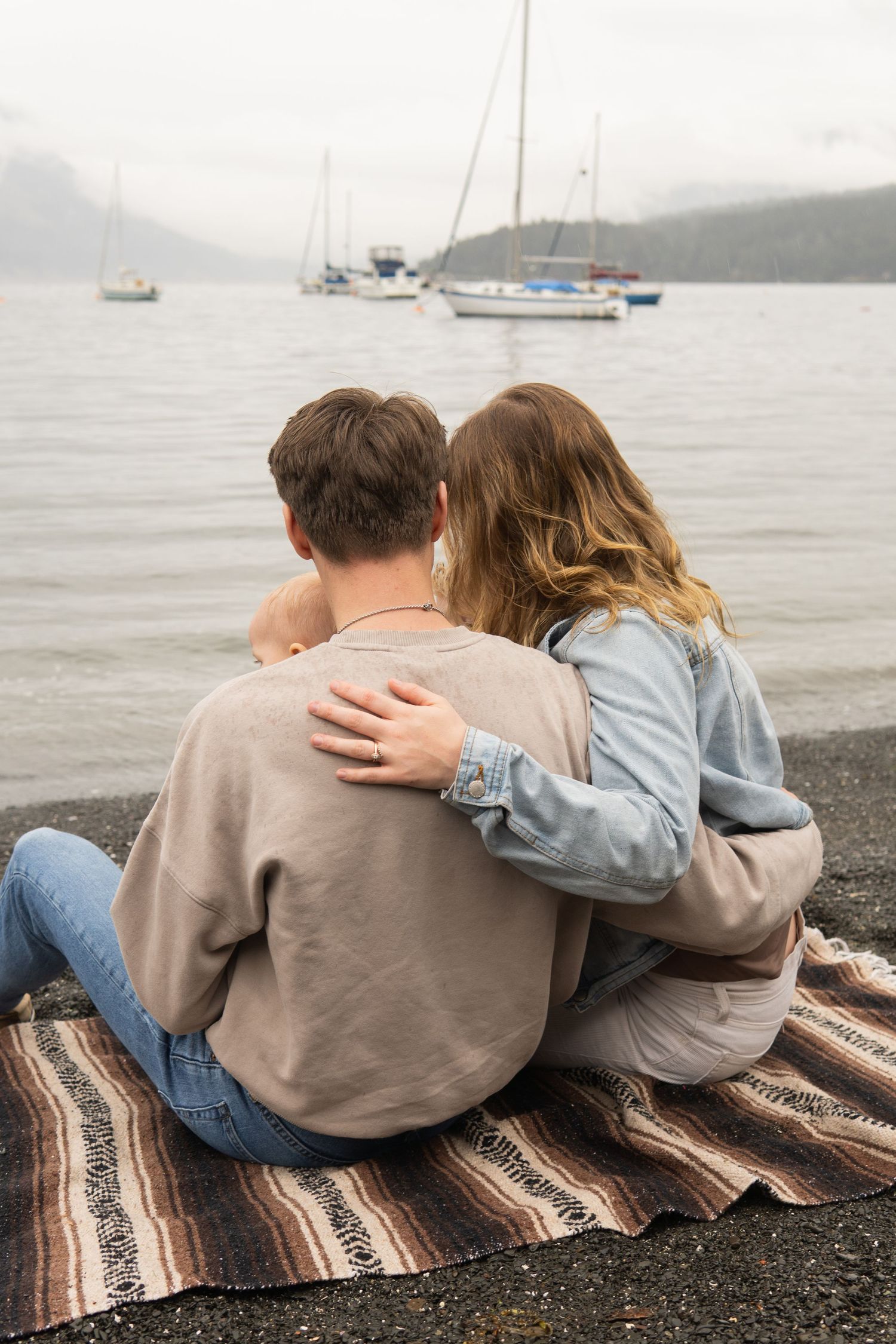 Couple sitting on a blanket by the water, embracing while watching sailboats on a misty bay with mountain backdrop.