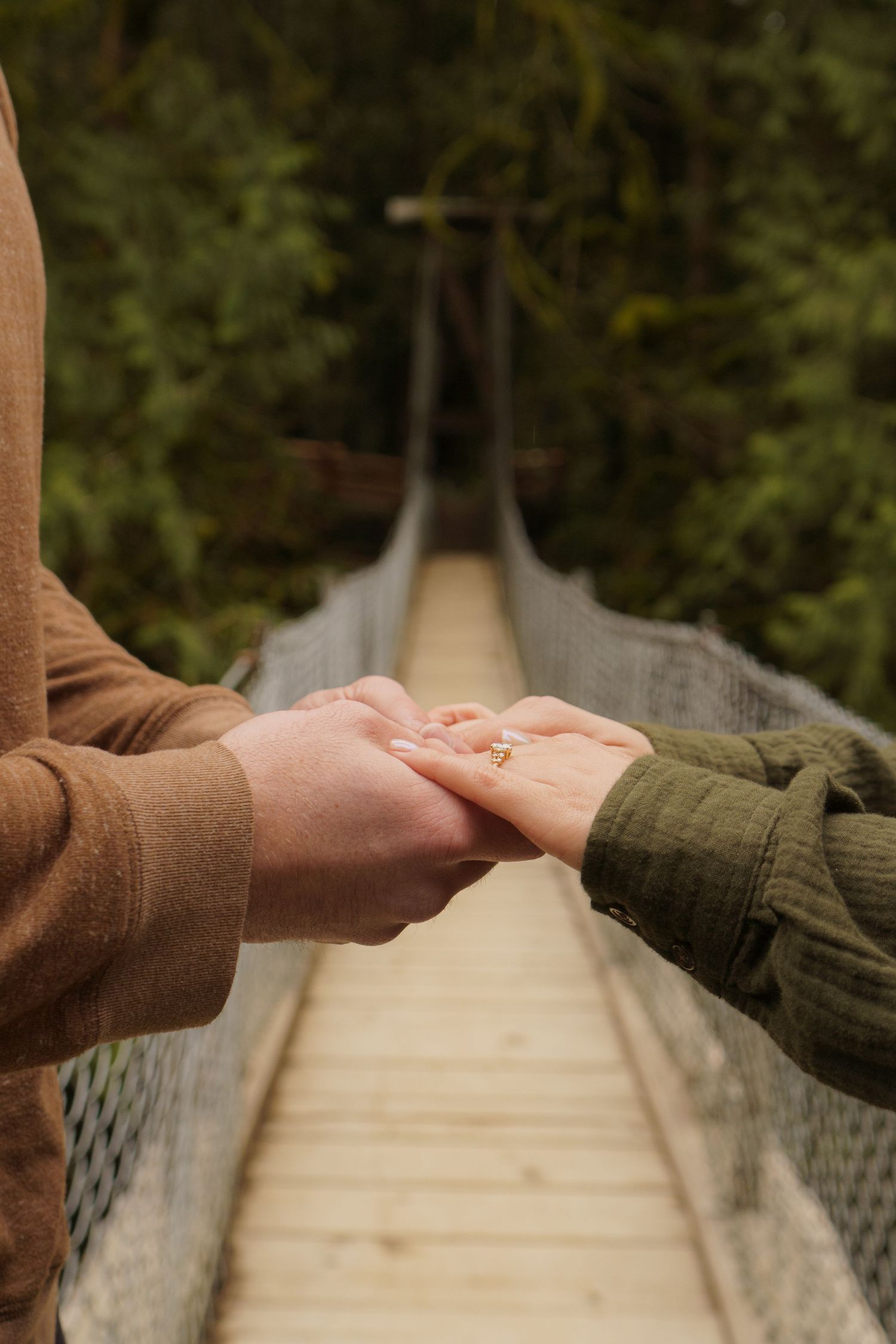 Close-up of couple holding hands showing engagement ring on a wooden suspension bridge.