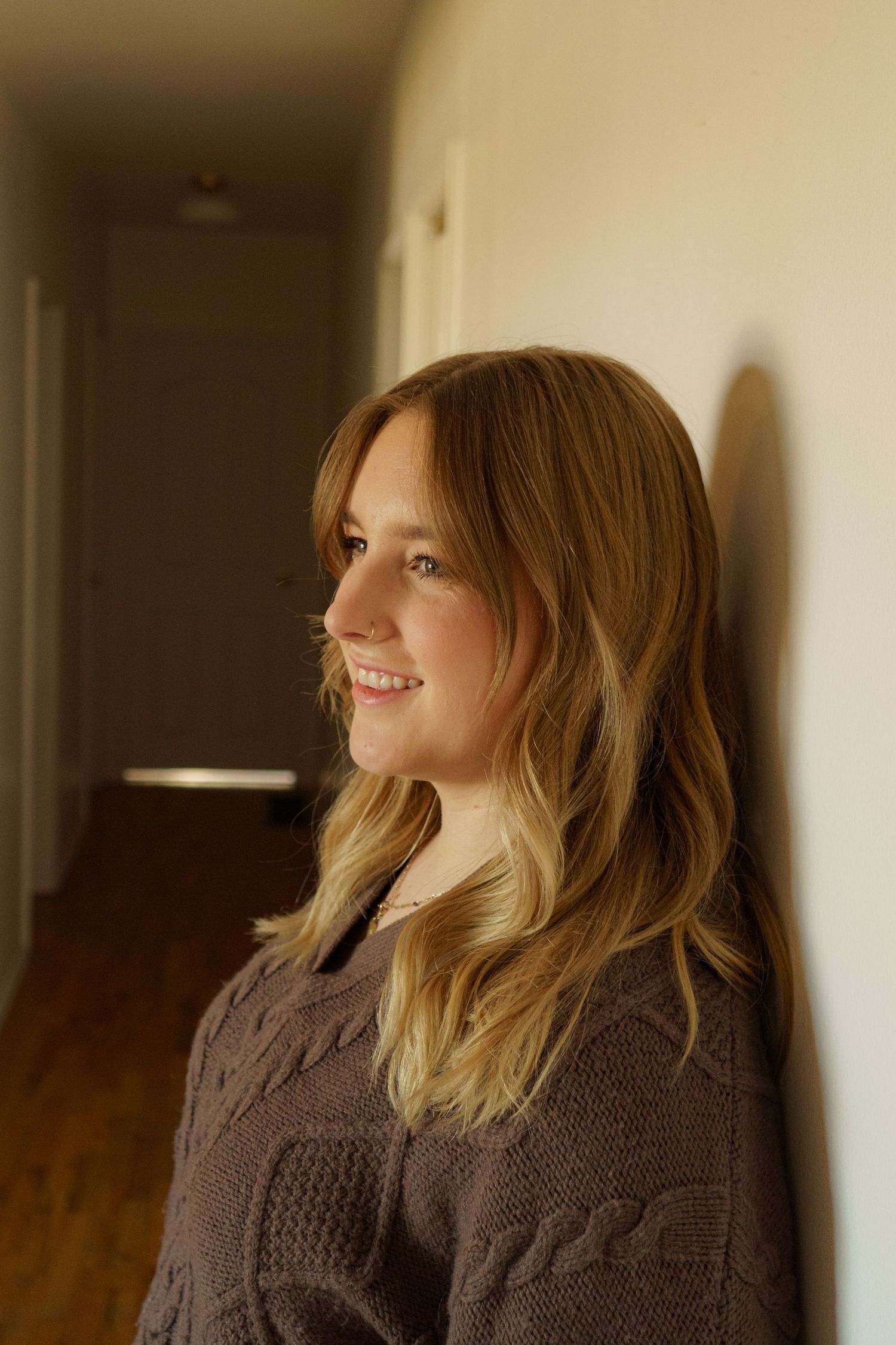 A young woman with blonde highlighted hair leans against a wall in a hallway, smiling softly in a brown knit sweater.