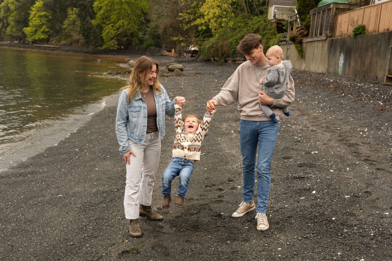 Family of four playing on a dark sandy beach, parents swinging young child between them.