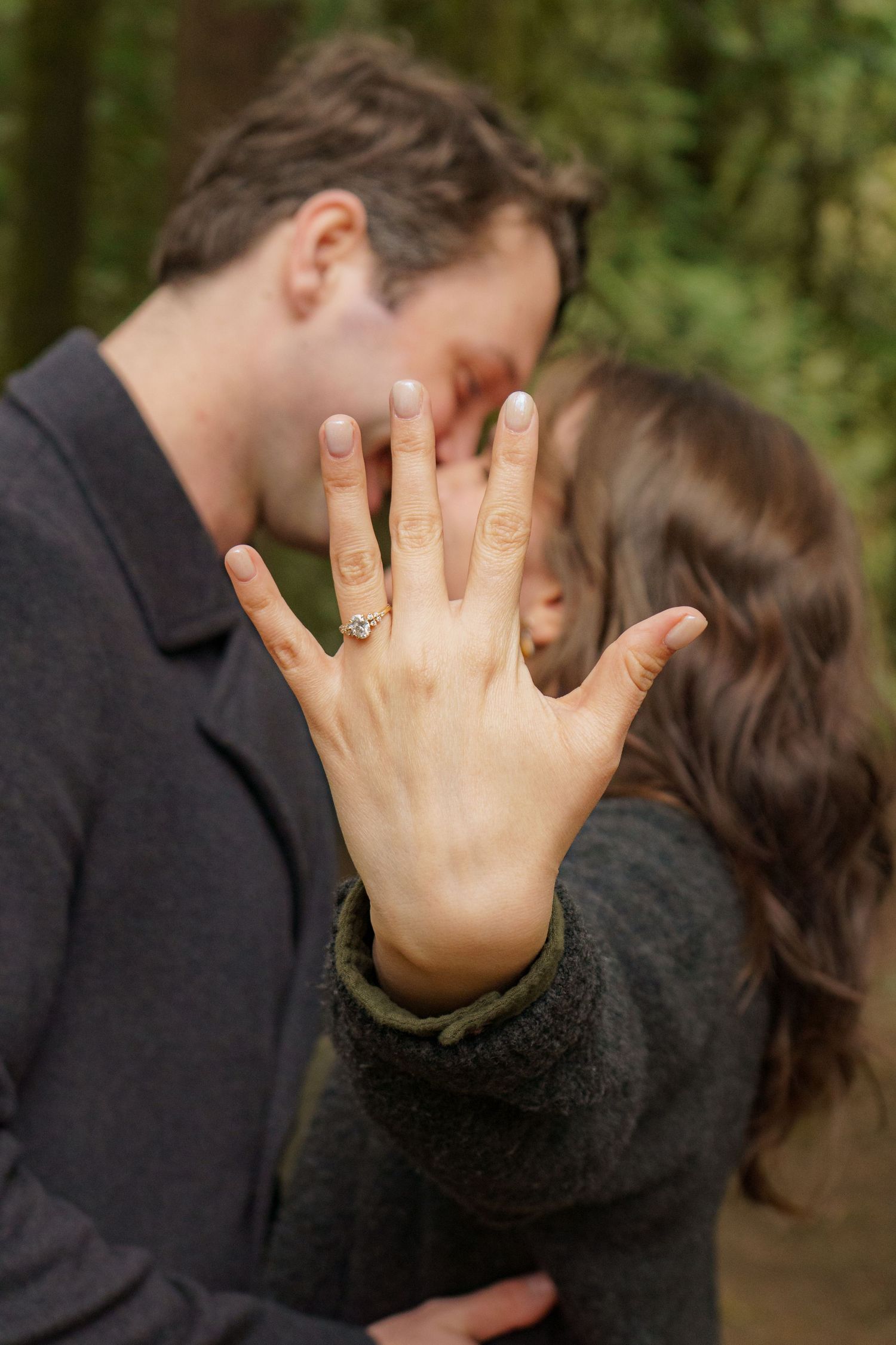 Couple kissing in forest while showing off engagement ring with diamond solitaire in focus foreground.