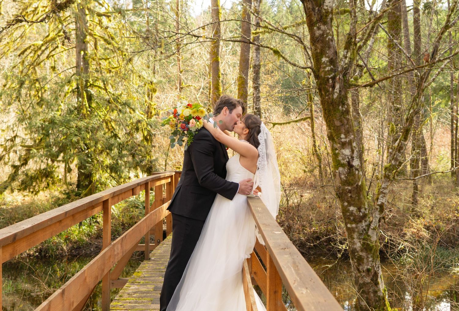 Bride and groom sharing a romantic kiss on a wooden bridge surrounded by lush greenery.