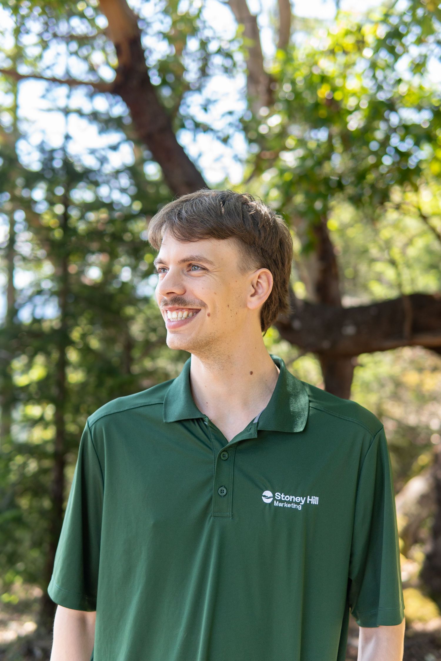 Smiling young man in green Stoney Hill polo shirt standing outdoors among trees on a sunny day.