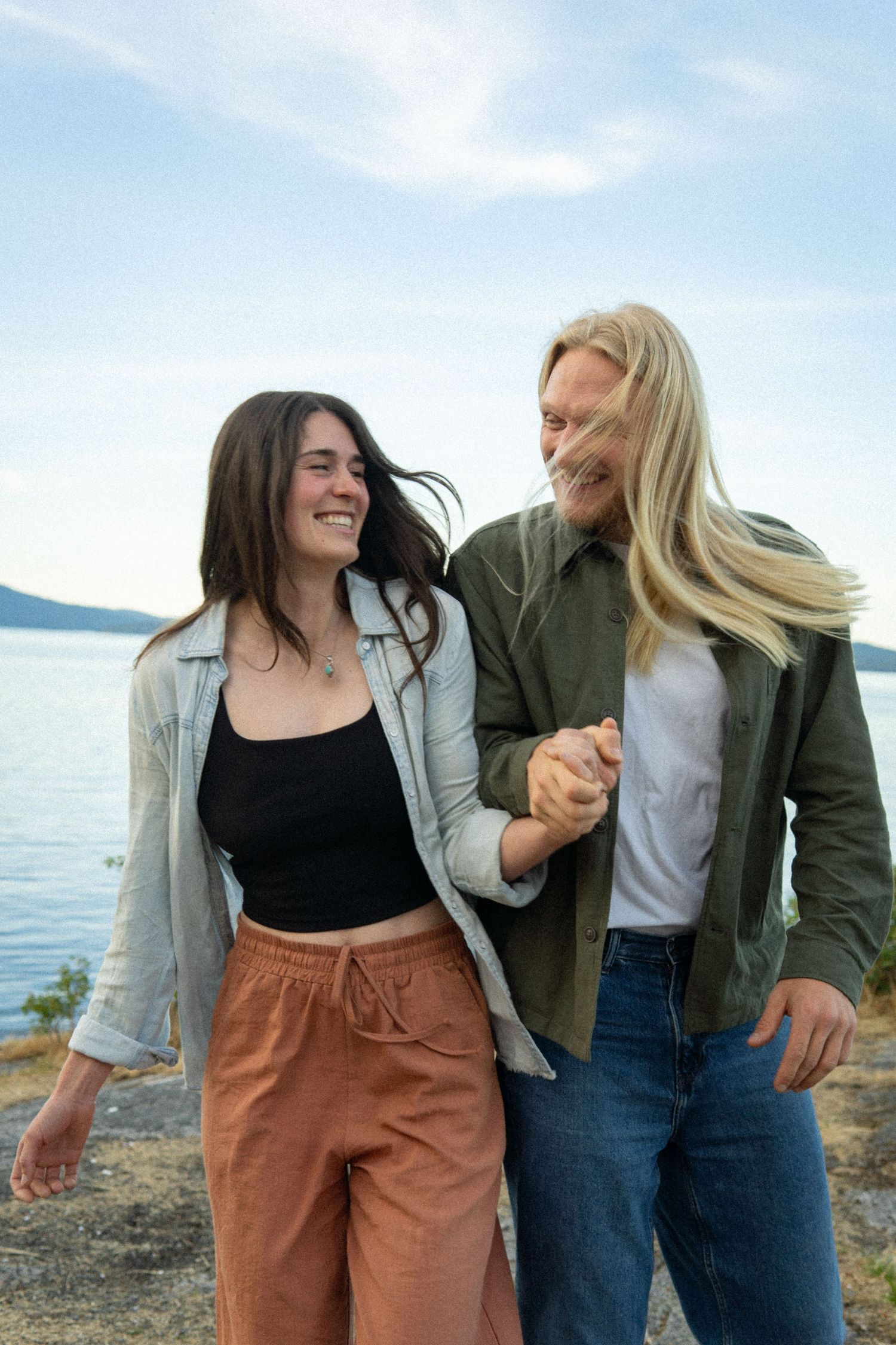 Smiling couple holding hands outdoors by a lake, woman in black top and orange pants, man in green shirt and jeans.