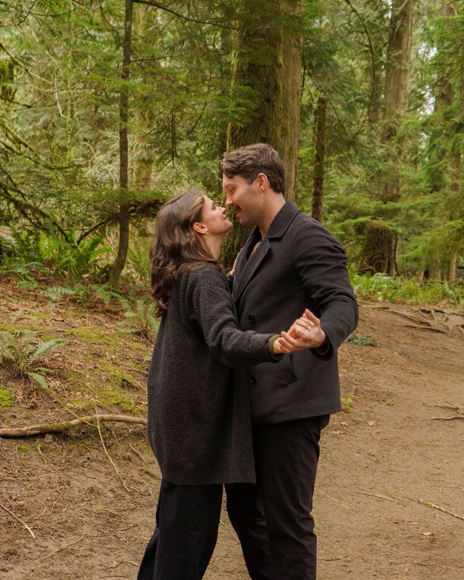 Couple sharing a romantic moment in a lush green forest, dressed in dark clothing, about to kiss on a dirt path.
