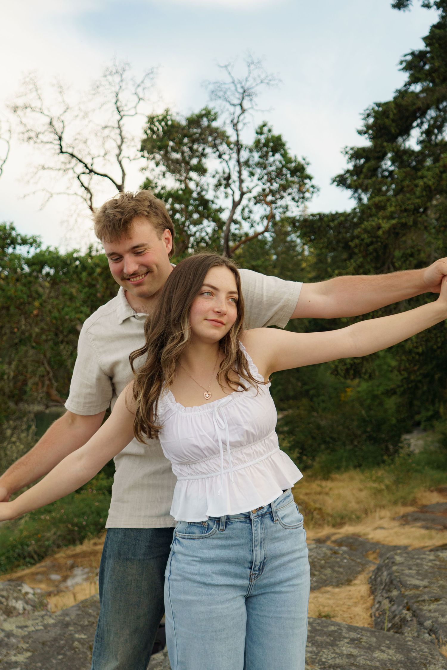 A smiling couple poses outdoors, woman in white top and jeans with arms spread wide, man behind her in neutral shirt.