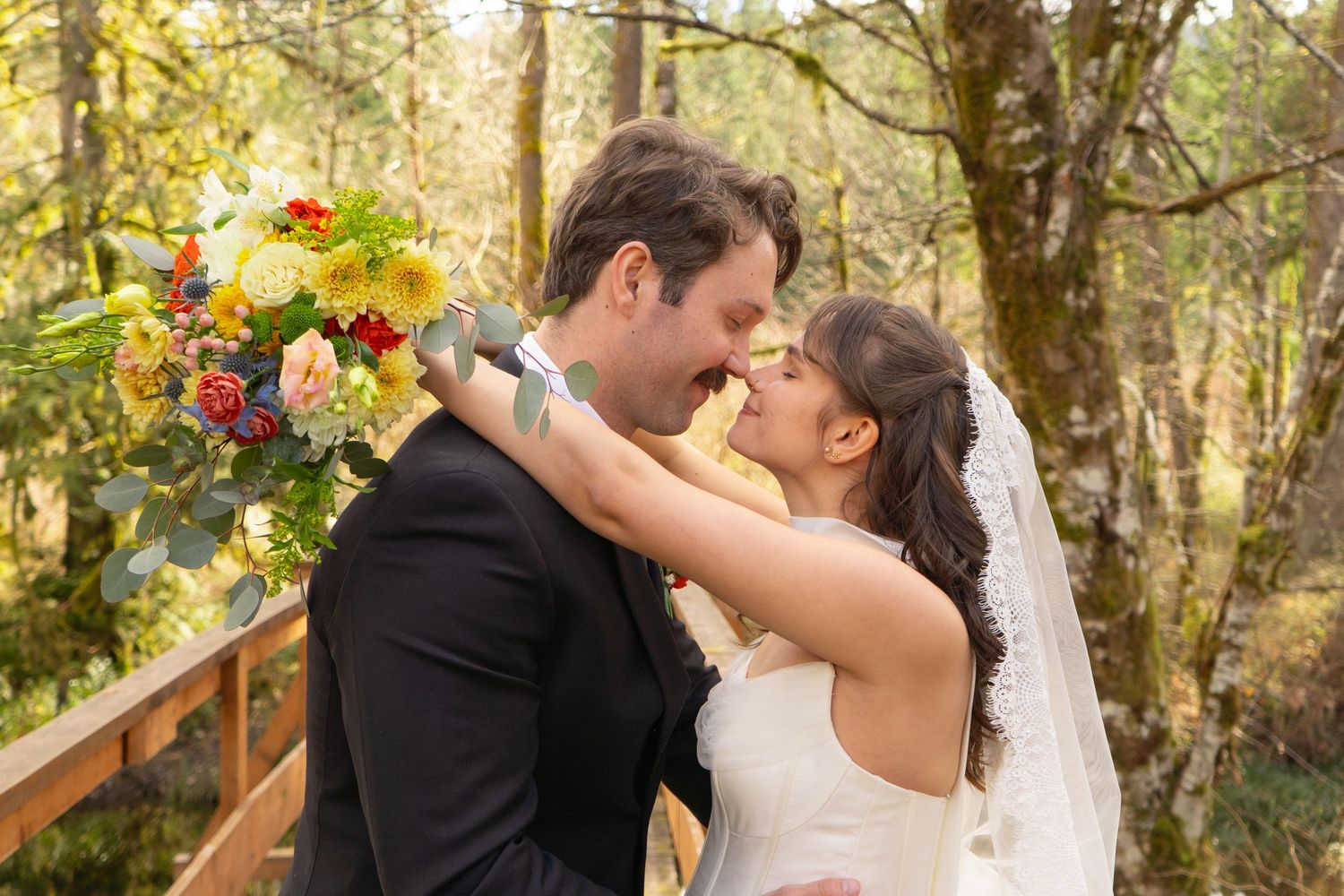 Bride and groom sharing a romantic kiss outdoors, bride holding colorful bouquet with red and yellow flowers.
