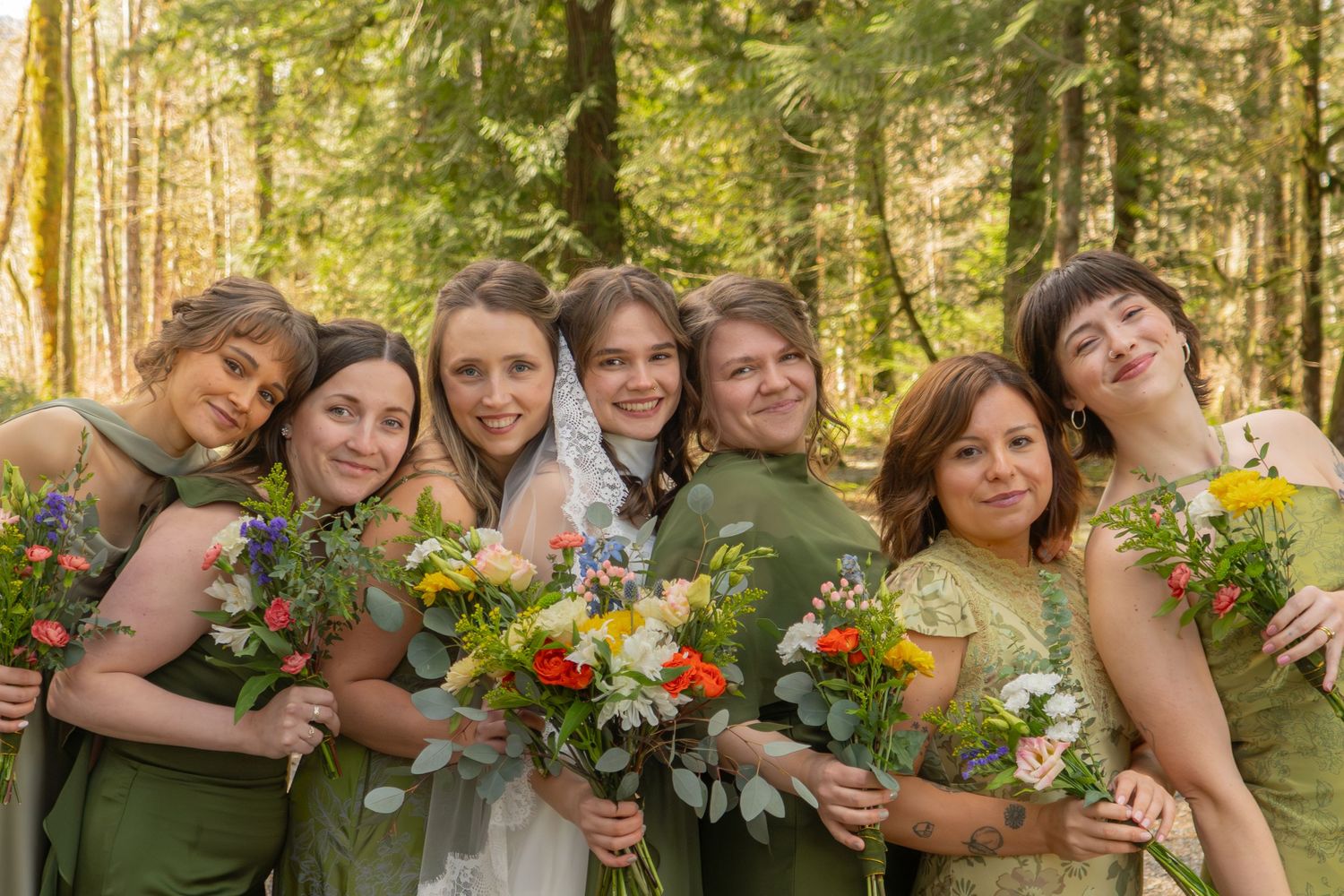 Bride in white veil with six bridesmaids in sage green dresses holding wildflower bouquets in a sunlit forest setting.