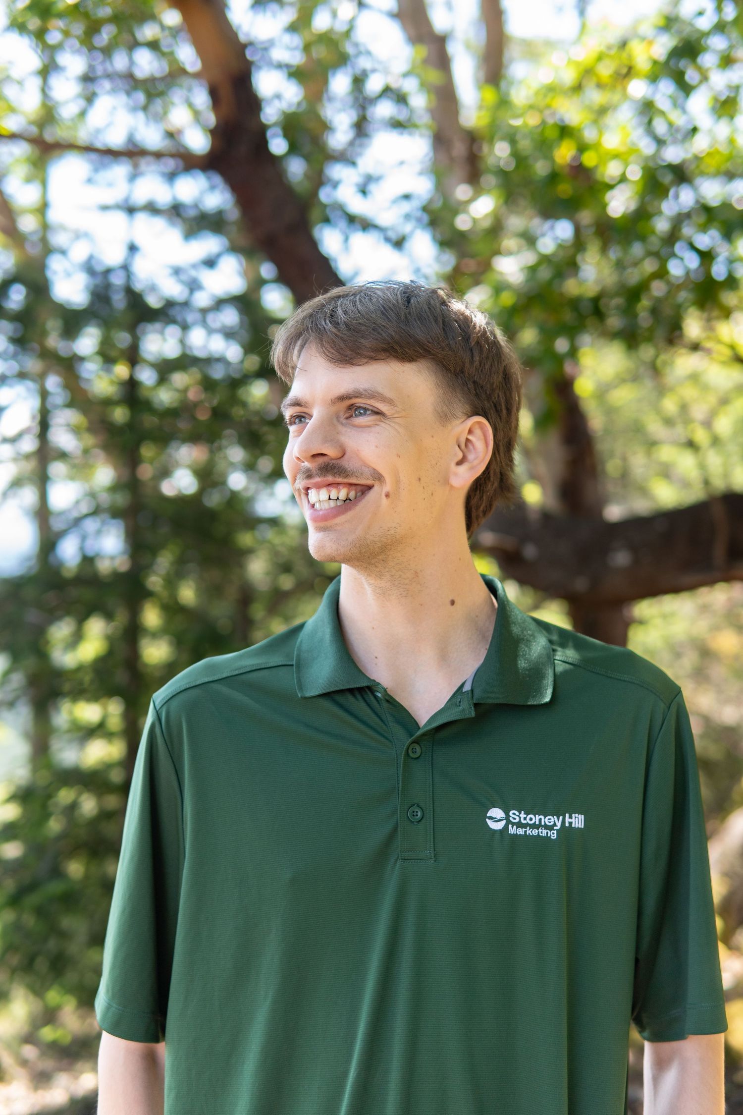 Smiling young man in a green Stoney Hill Brewing polo shirt outdoors with trees in the background.