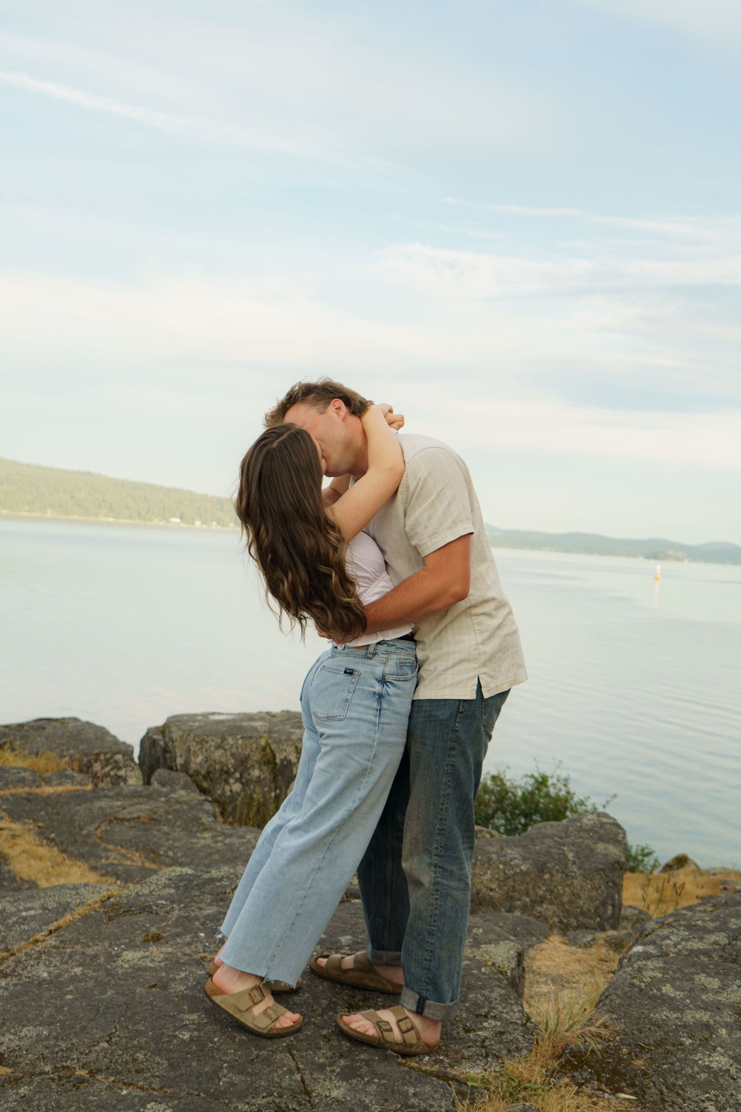 Couple kissing on rocky shoreline with calm blue water and hills in background, wearing jeans and casual tops.