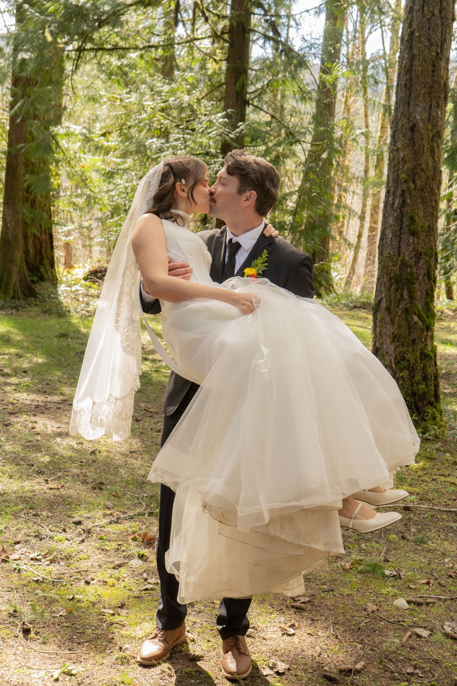Groom carries bride in white wedding dress and veil through sunlit forest, sharing a romantic kiss.