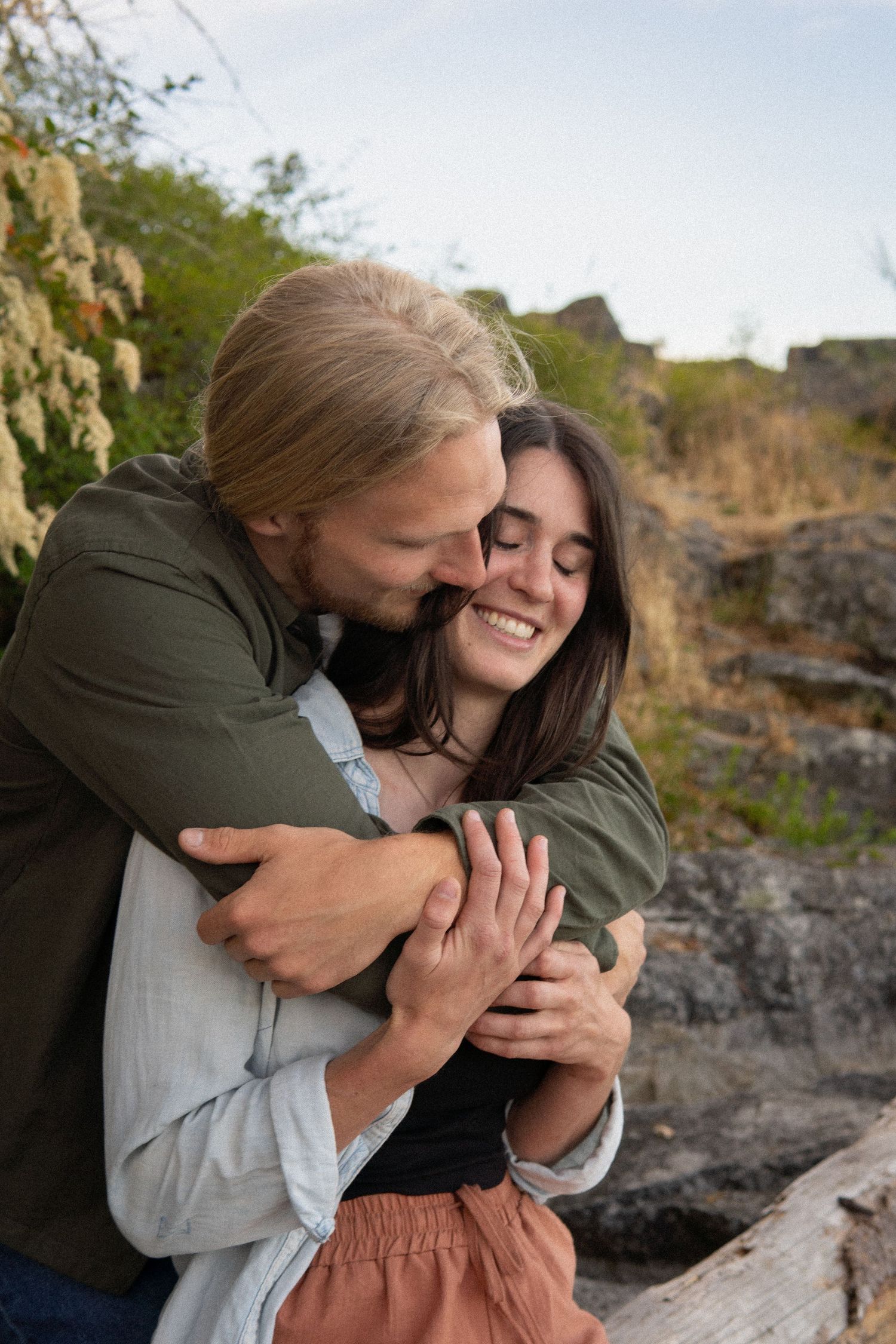A smiling couple embracing warmly outdoors near rocky terrain and wild vegetation.