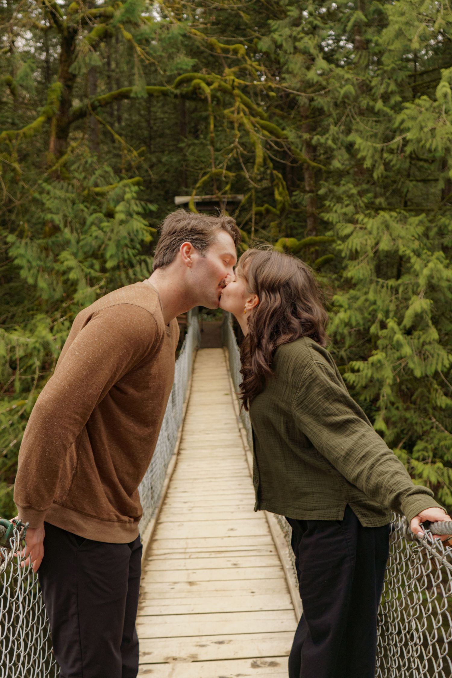 Couple sharing a kiss on a wooden suspension bridge surrounded by green forest trees.
