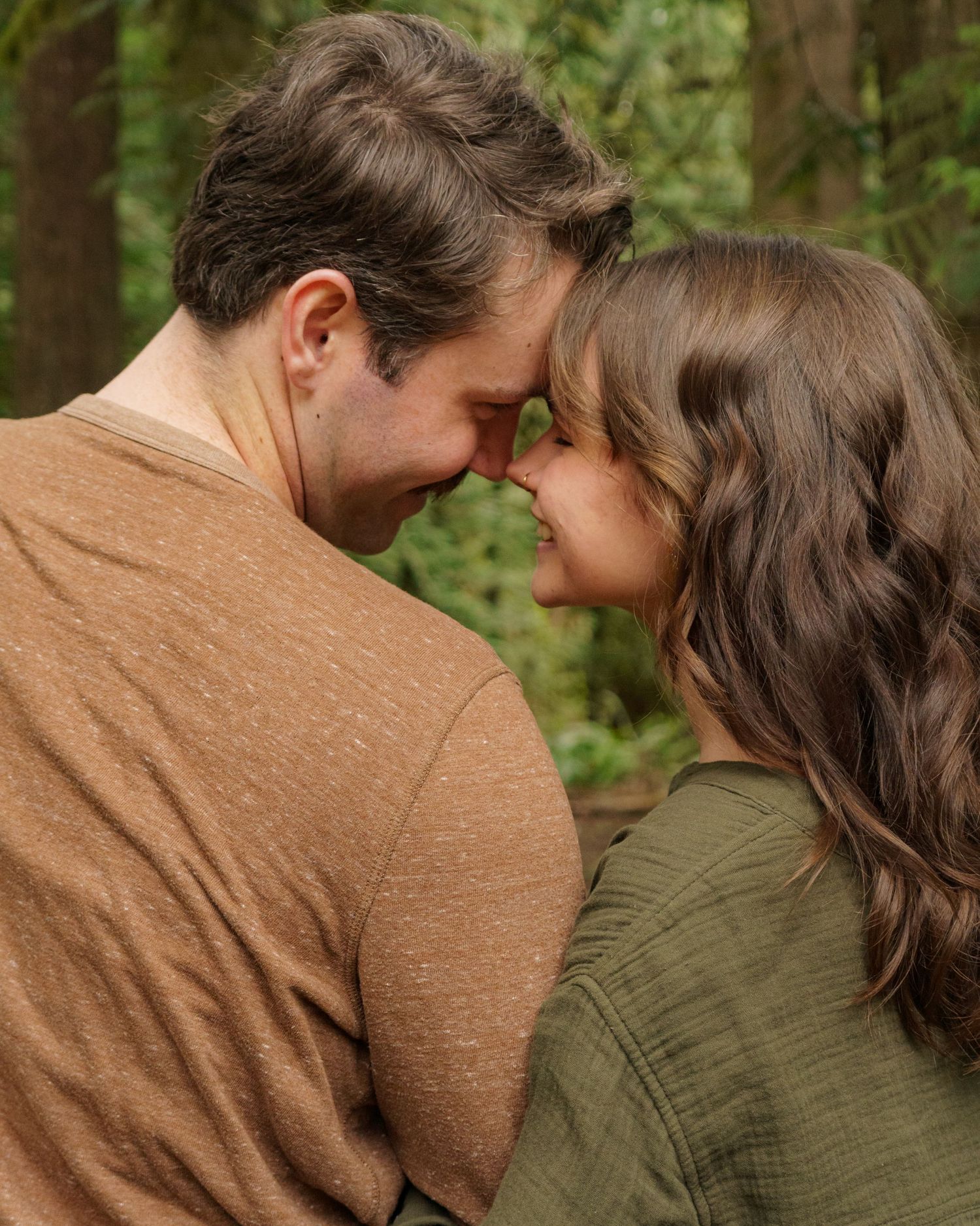 A romantic couple touching foreheads in a lush green forest, sharing an intimate moment outdoors.