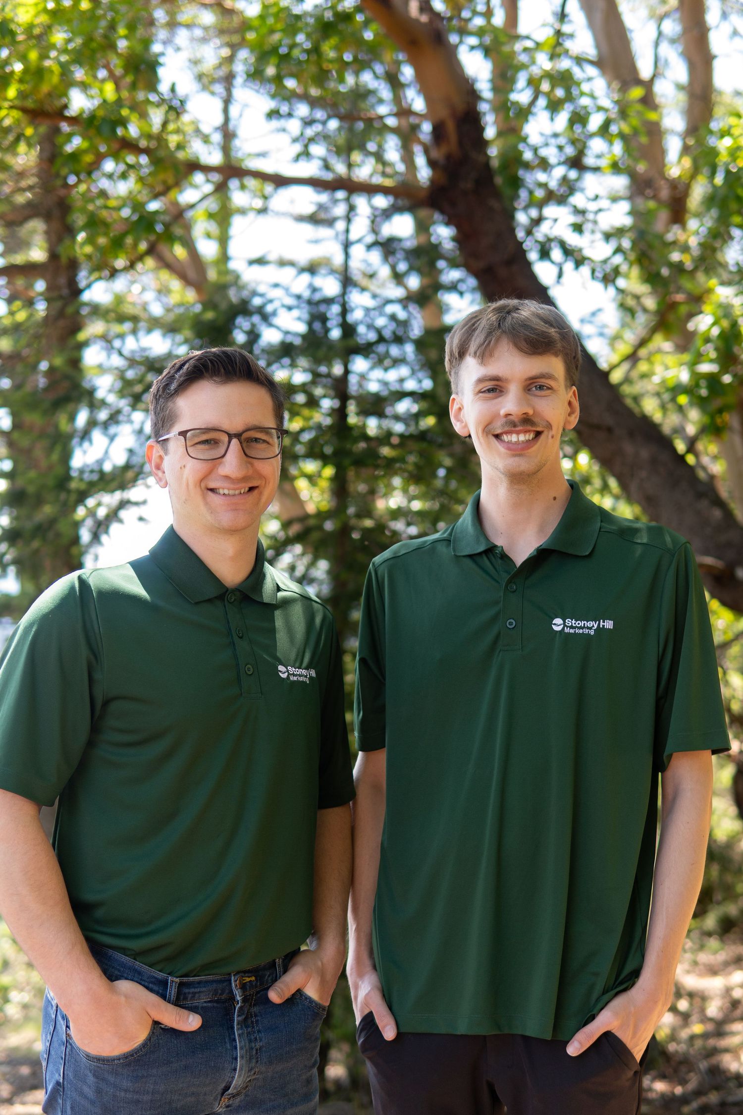 Two smiling men in matching green polo shirts stand outdoors among trees in a professional photo.