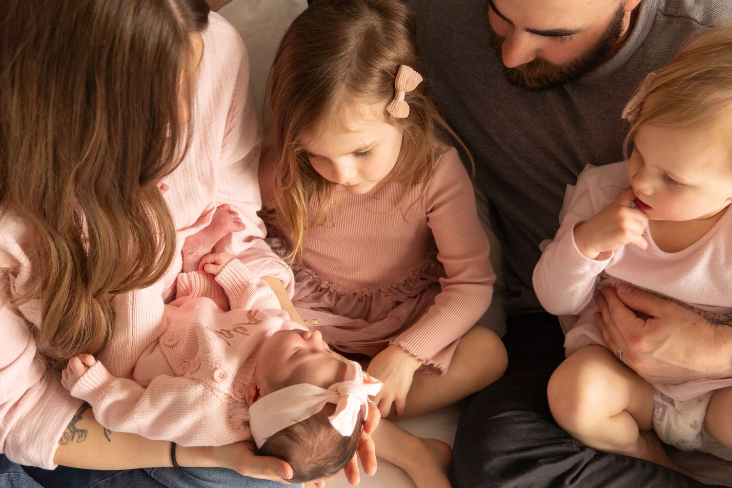Family gathered closely admiring a newborn baby, with two young girls in pink dresses and parents sharing a tender moment.