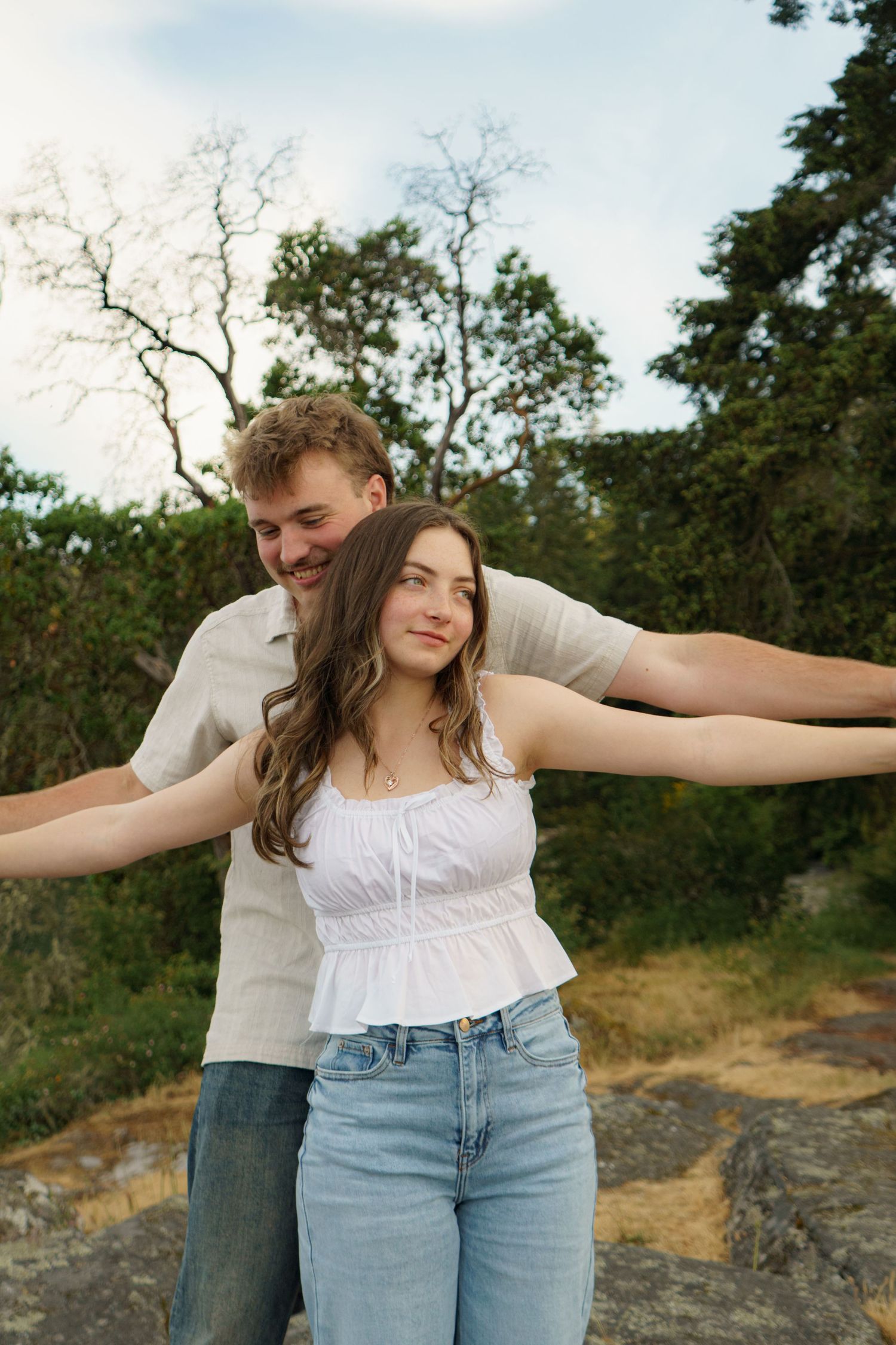 A young couple poses outdoors, woman in white top and jeans spreads her arms wide while man smiles behind her.