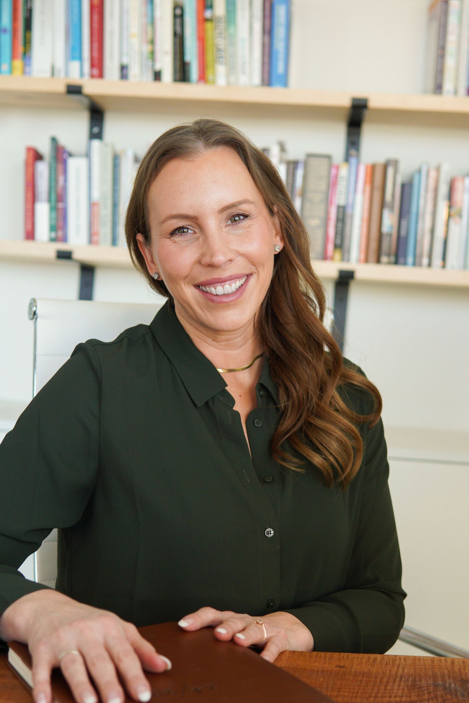 Smiling woman in green blouse seated at desk with bookshelf background in professional office setting.
