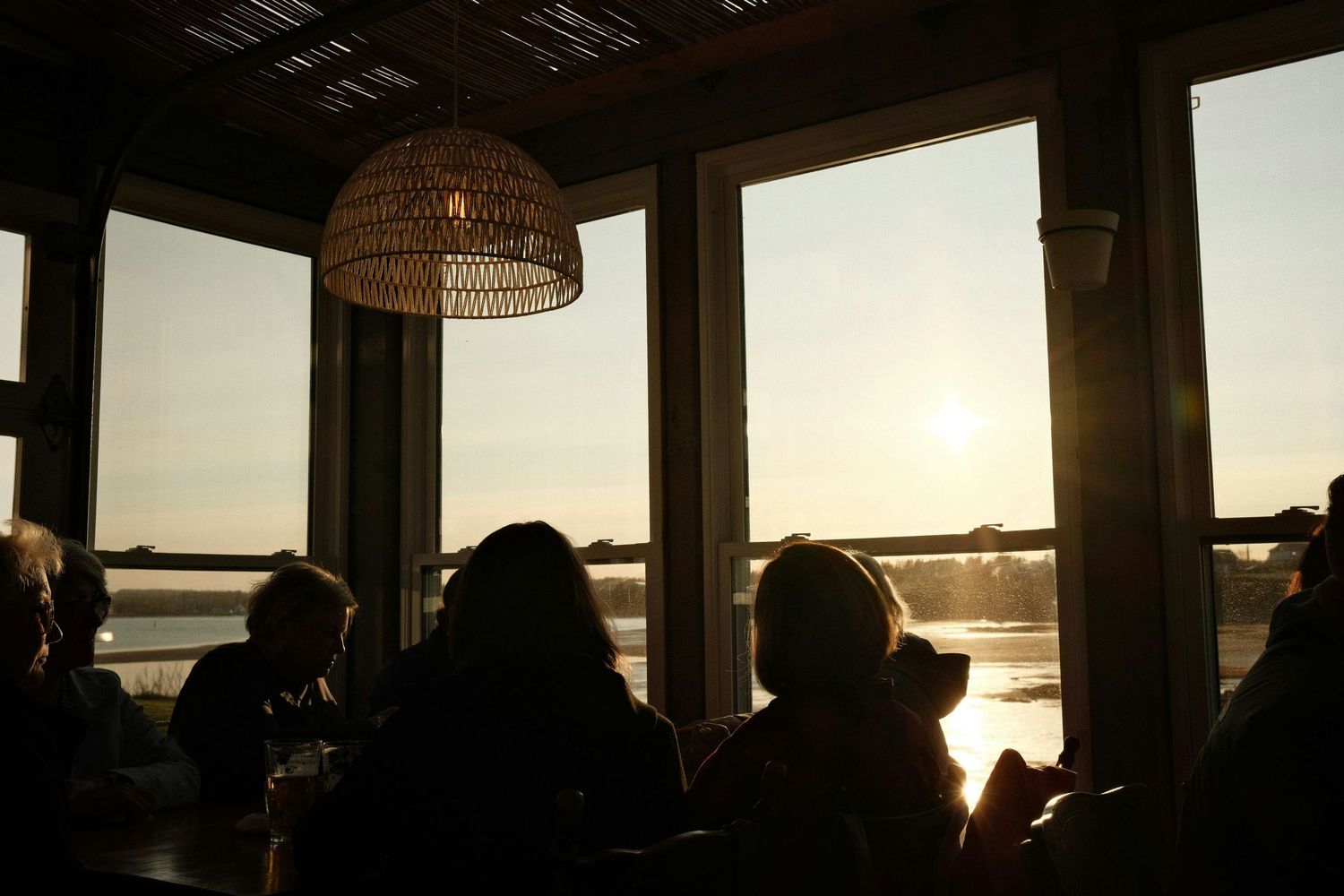 Silhouettes of people dining by large windows with a stunning sunset over water view.
