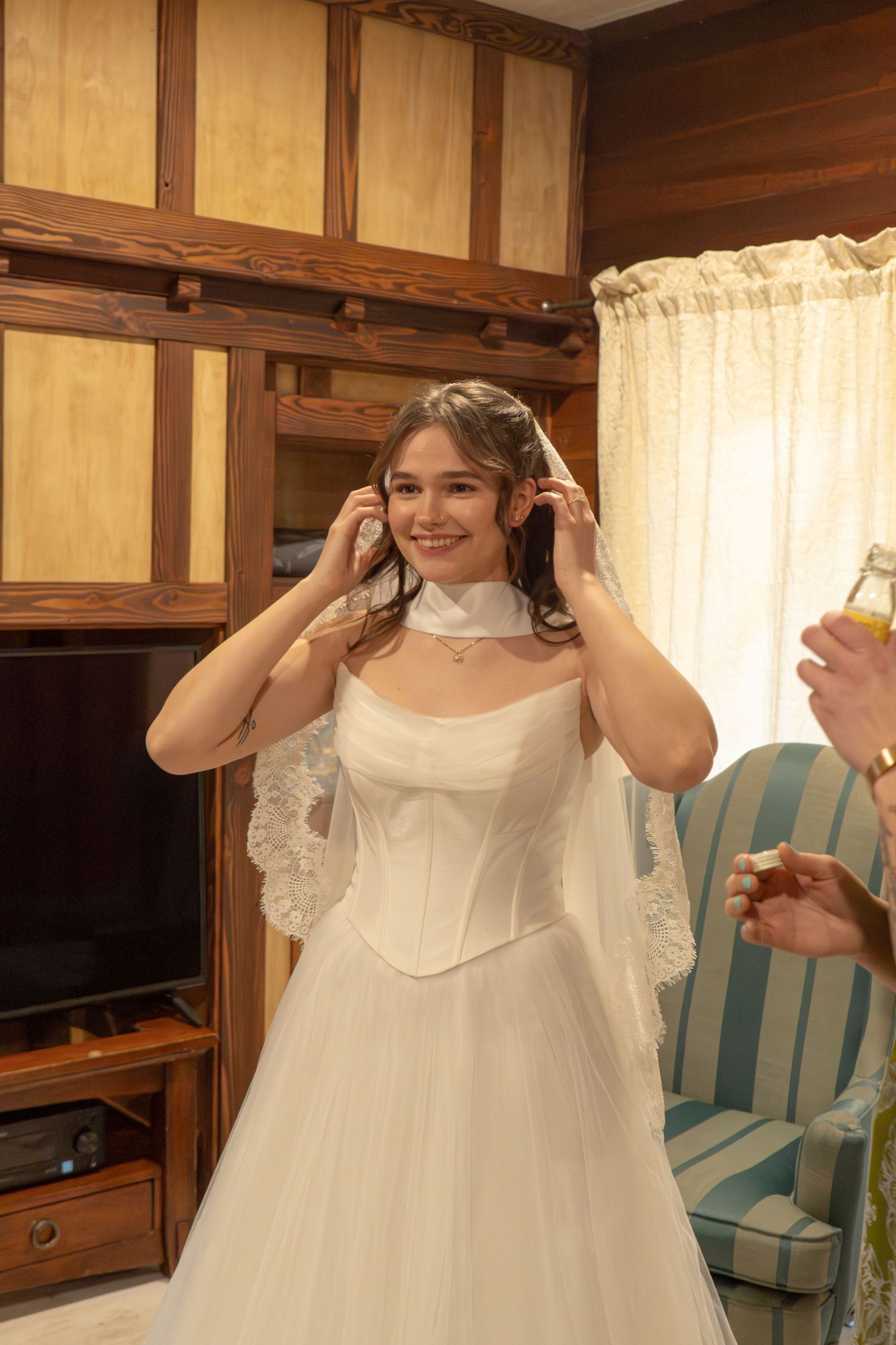 Smiling bride in white corset wedding dress adjusting her lace veil in a wood-paneled room.