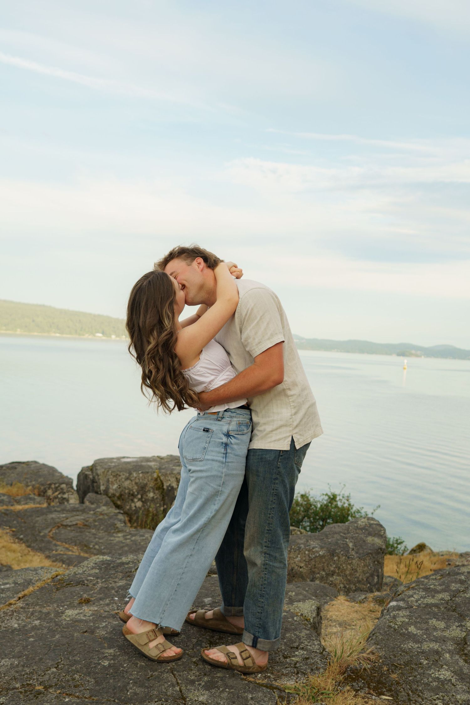 Couple sharing a romantic kiss on rocky waterfront with scenic lake and mountains in the background.