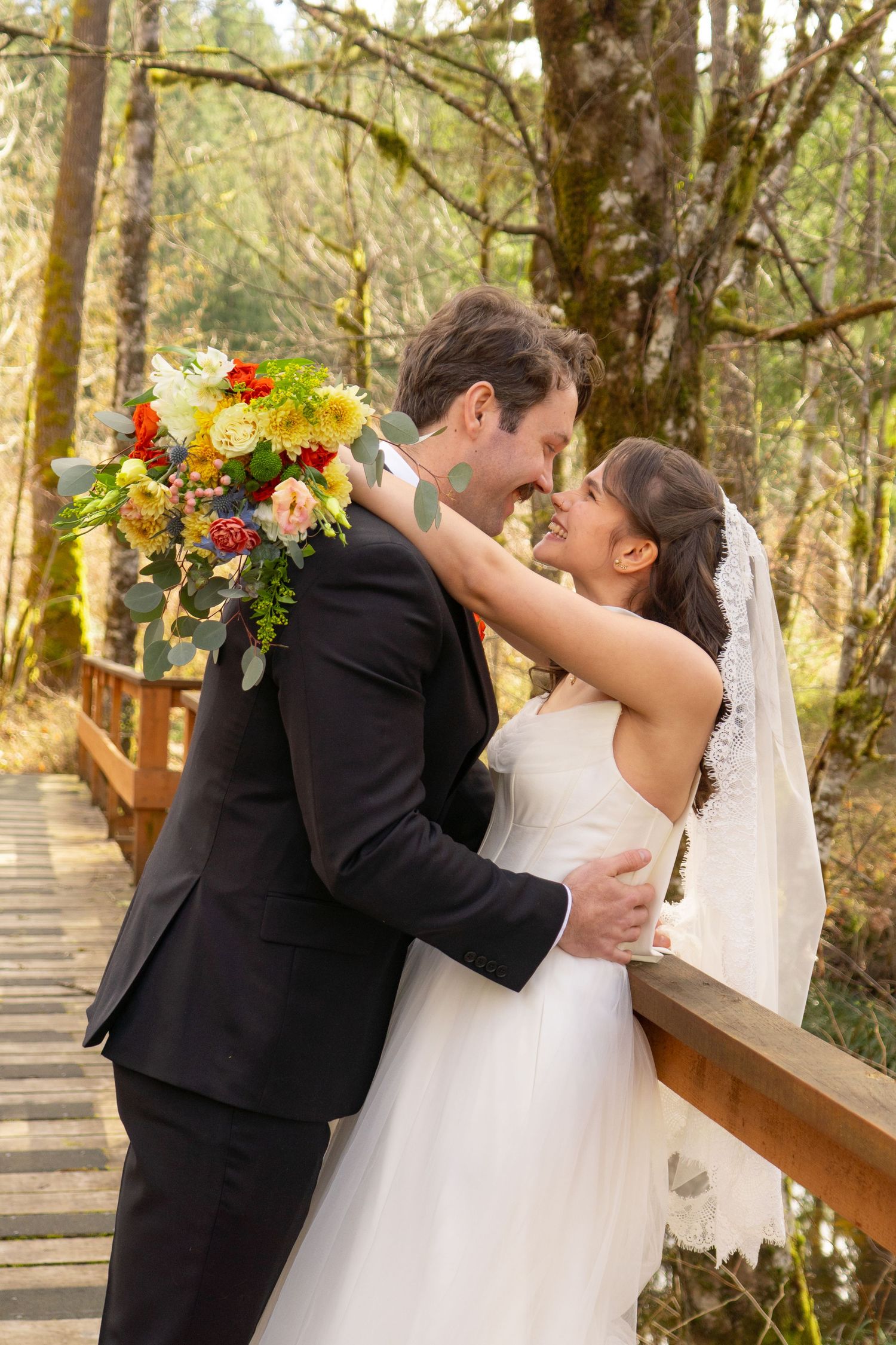 Bride and groom nearly kissing on wooden bridge with colorful fall bouquet.