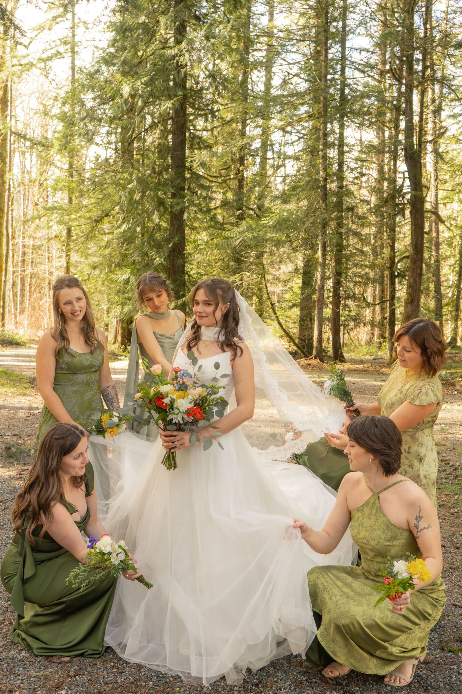 Bridesmaids in green dresses helping bride arrange her veil and bouquet in a sunny forest setting.