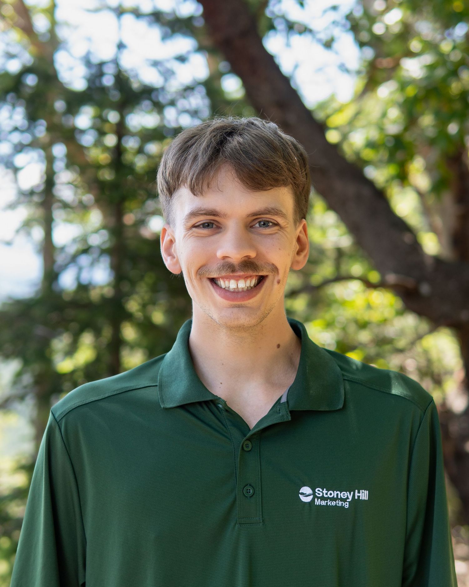 Young man smiling outdoors in a green Stoney Hill Mentoring polo shirt with trees in the background.