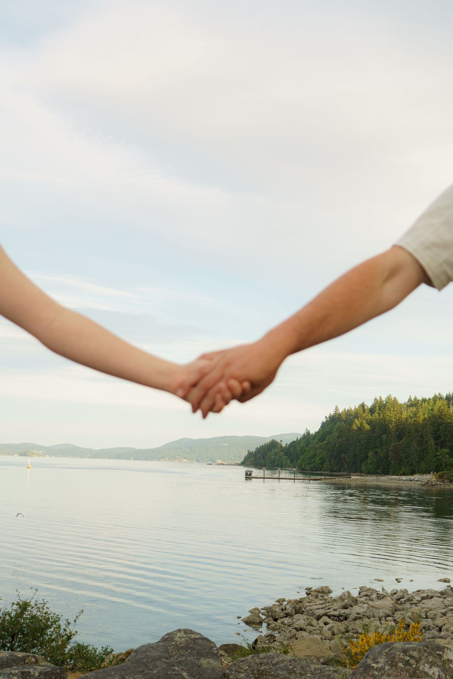 Couple holding hands by a serene lake with forested hills and a dock in the background.