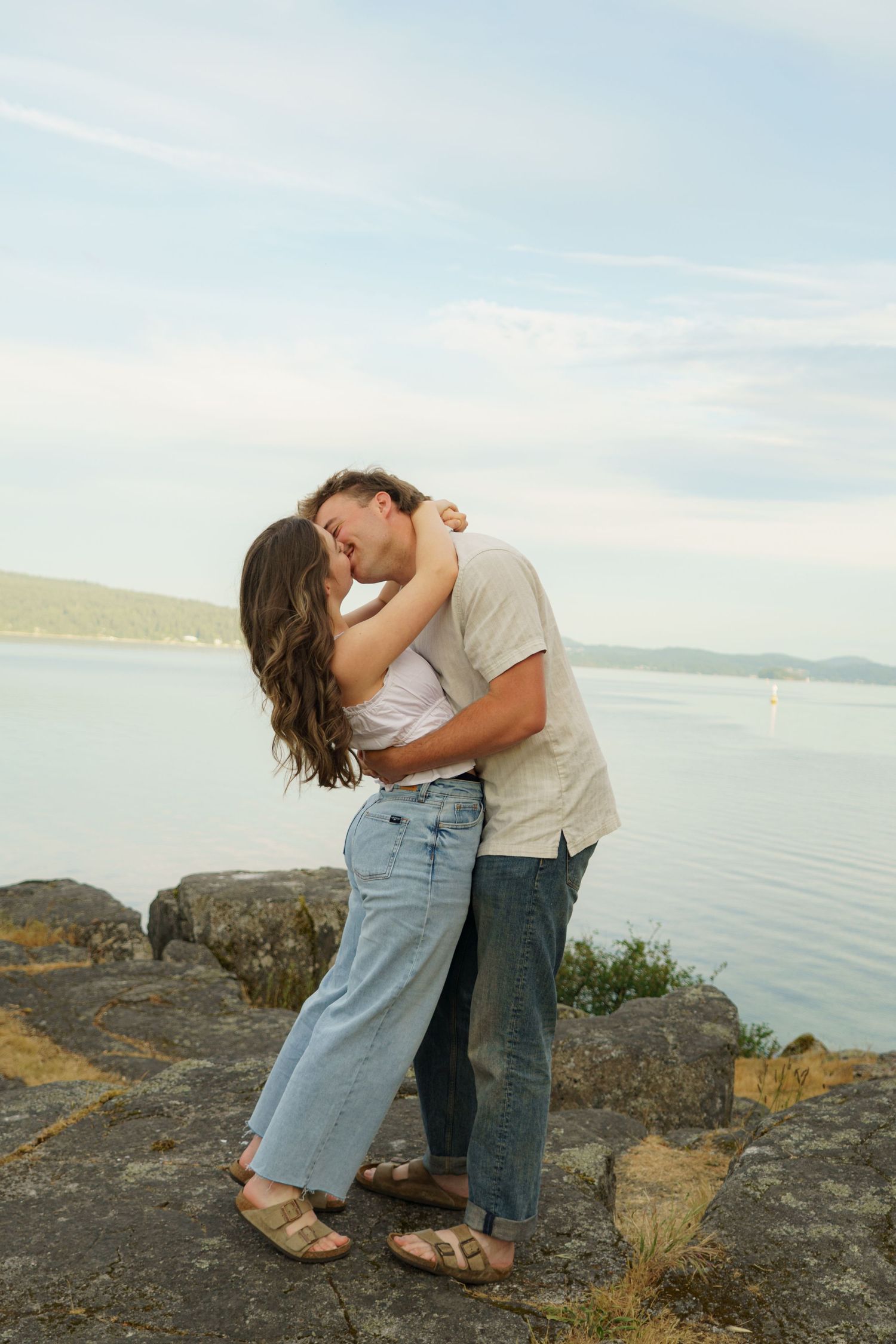 Couple sharing a romantic kiss on rocky waterfront with scenic lake and mountains in the background.