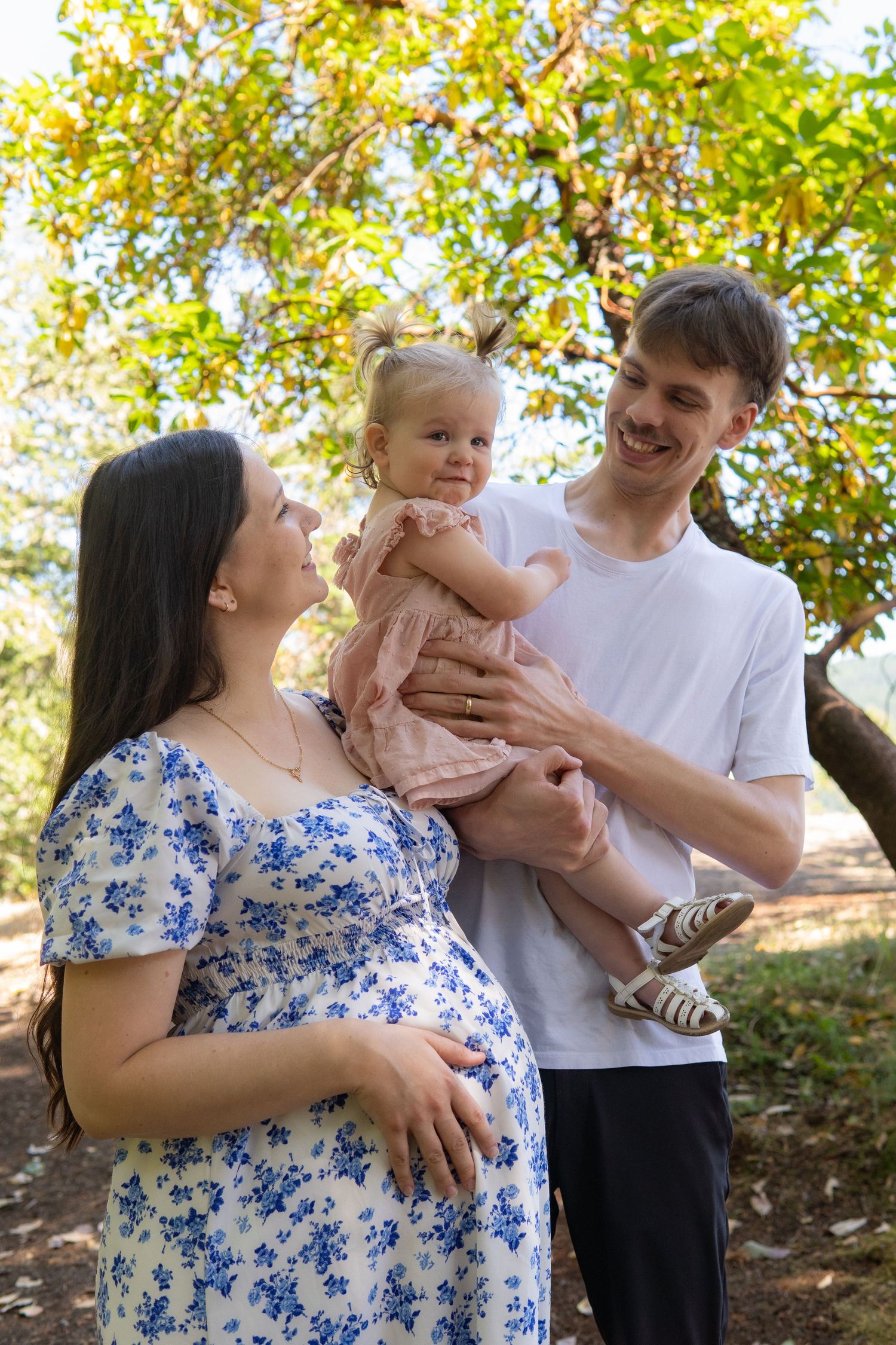 Happy pregnant woman in floral dress with husband holding toddler daughter outdoors in sunny park.