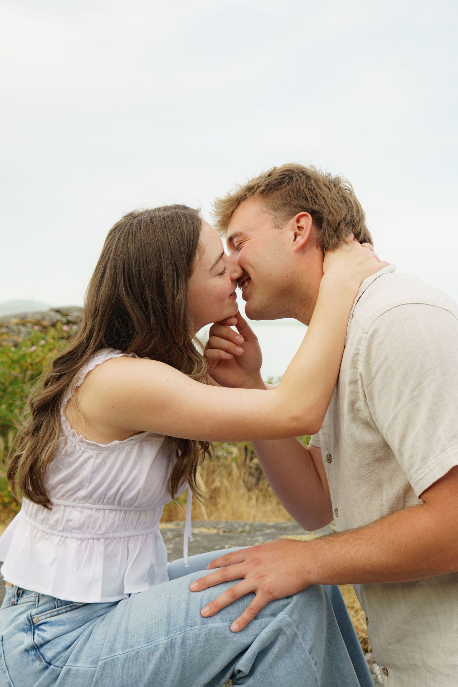 A young couple sharing a romantic kiss outdoors, sitting together against a natural countryside backdrop.