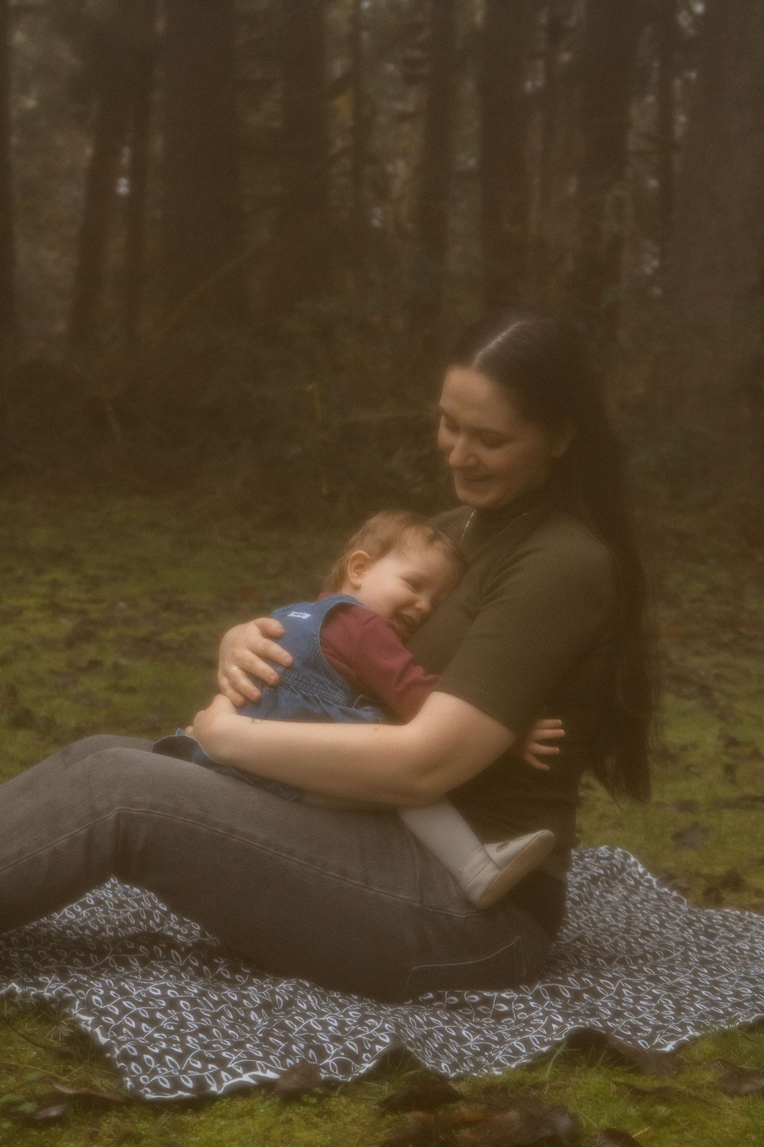 A mother tenderly holds her sleeping baby on a blanket in a misty forest setting.