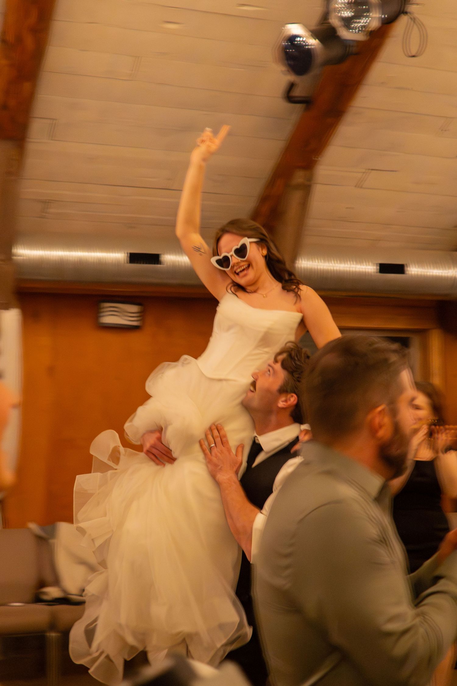 Excited bride in sunglasses raised by groom during energetic wedding reception celebration.