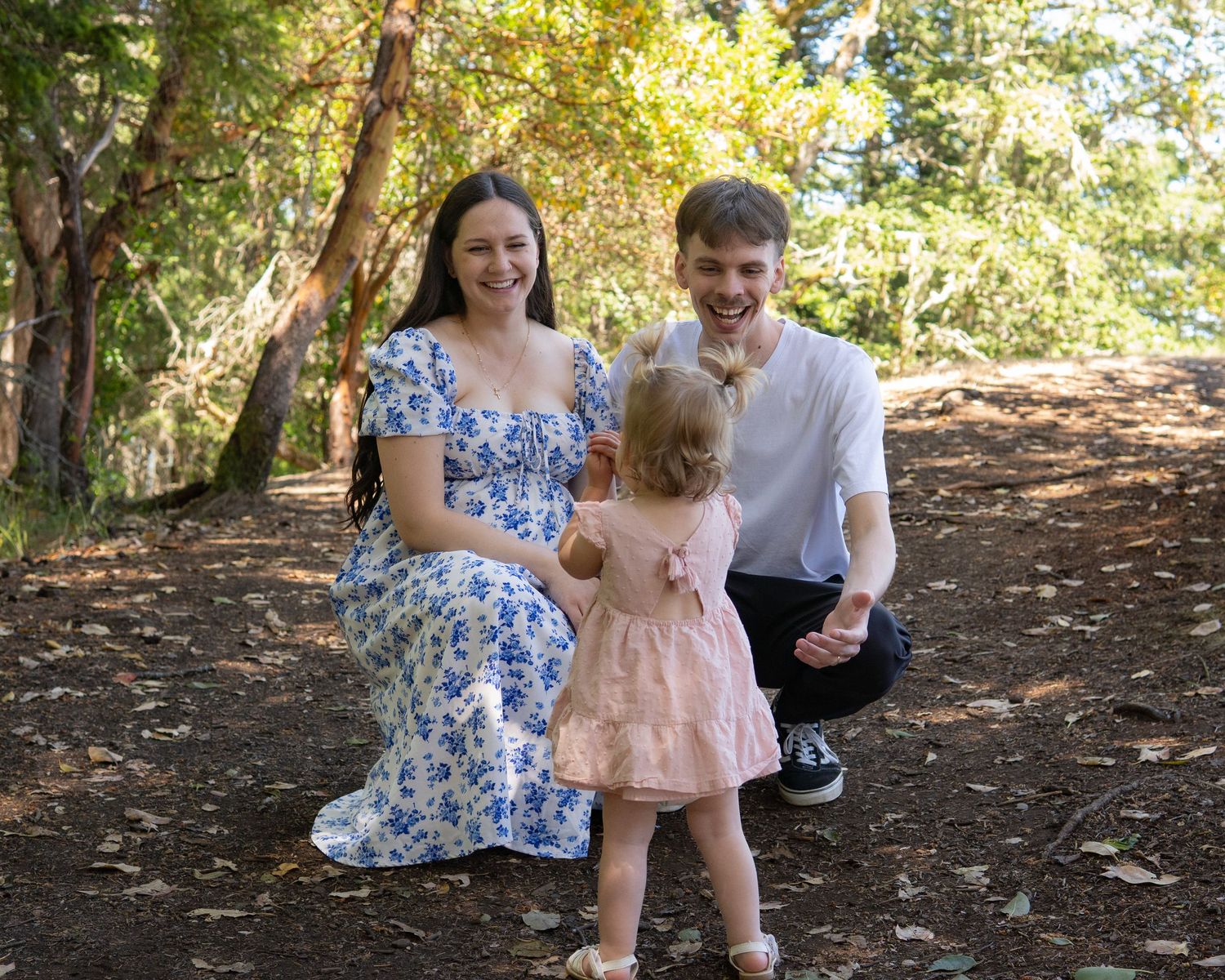 Happy young family with toddler daughter smiling outdoors in a sunlit forest setting.