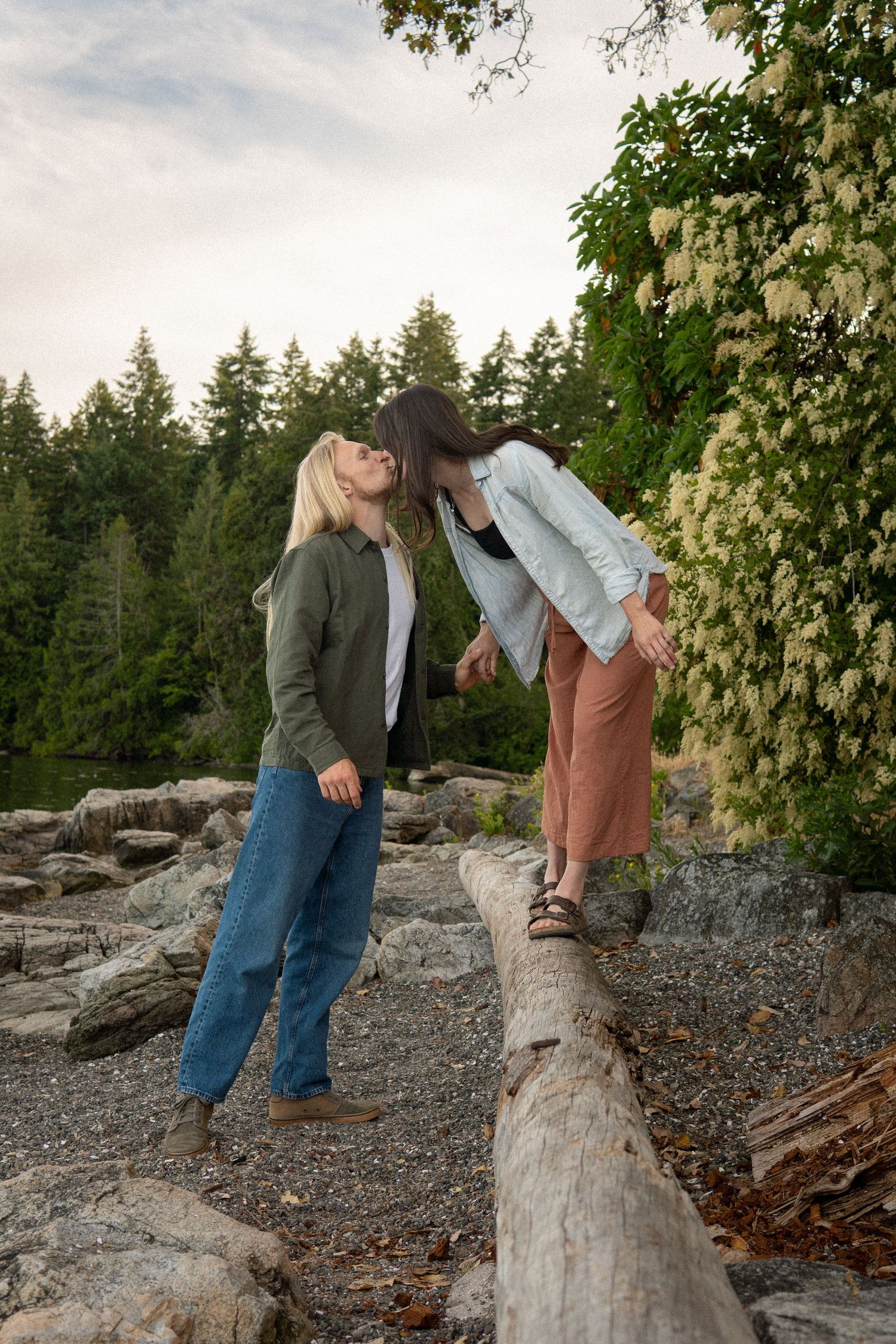 Two women sharing a kiss near a fallen log surrounded by rocks and lush greenery.